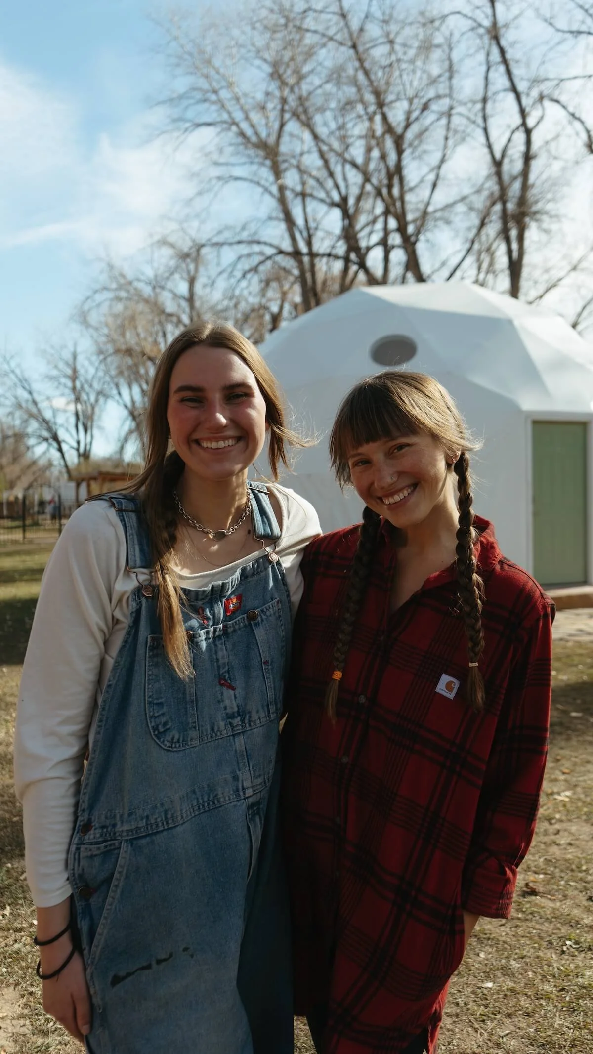 Two young women with long hair and braids smiling outdoors, standing in front of a white geodesic dome structure, with leafless trees and a clear sky in the background.