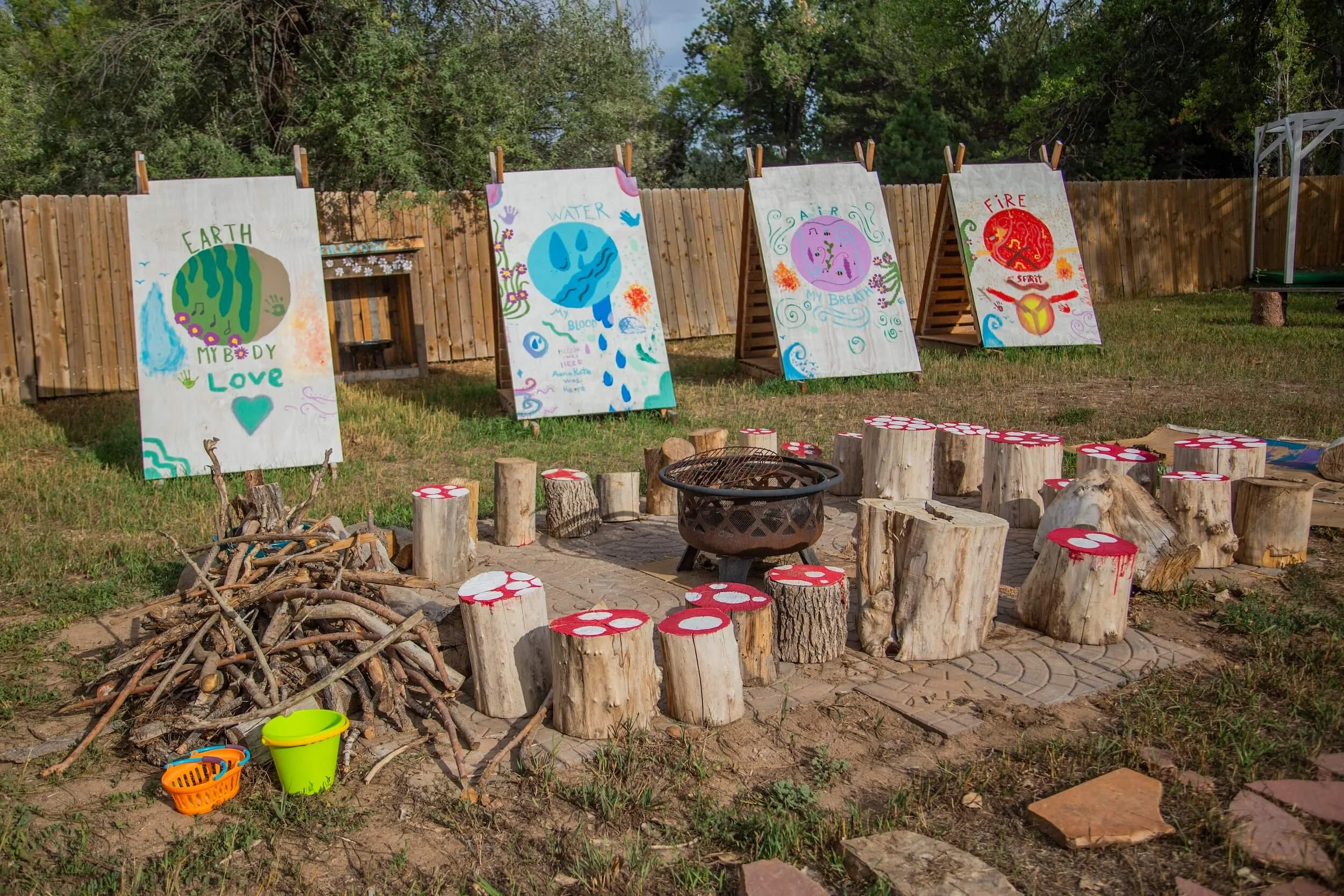 Outdoor art display with colorful paintings on large boards attached to a wooden fence, surrounded by tree trunks, branches, and plastic buckets, and a fire pit in foreground.