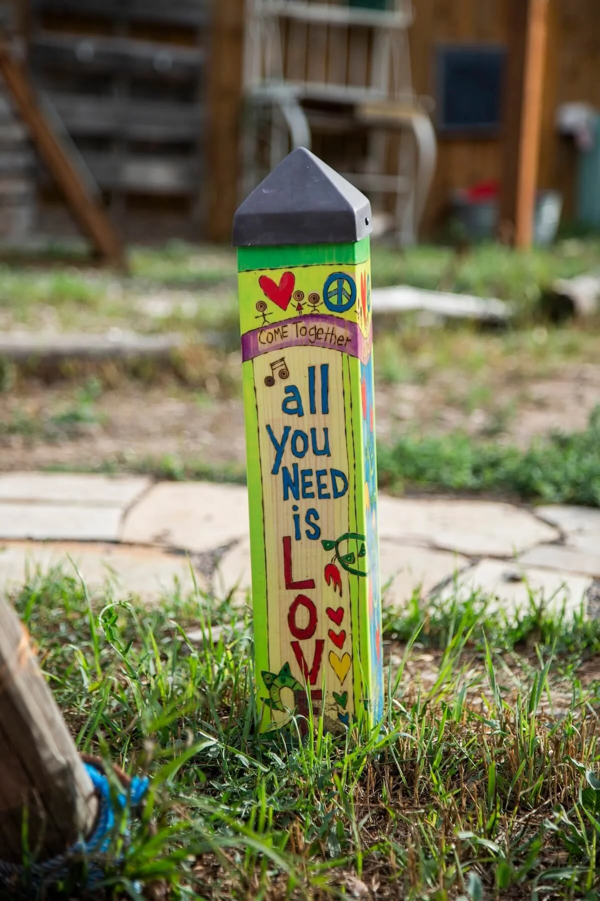 Colorfully painted wooden stake with messages of love, peace, and togetherness, decorated with hearts, peace signs, and smiley faces in a grassy yard.