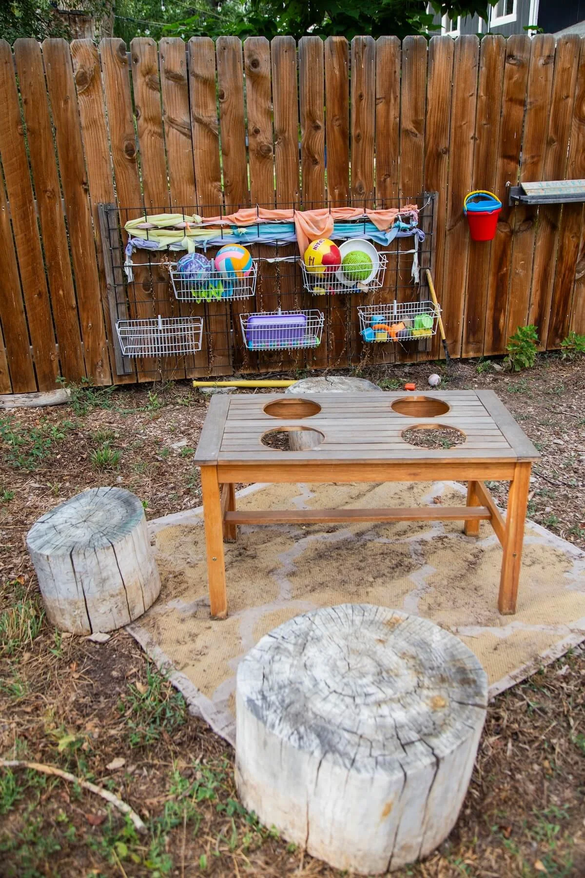Backyard space with a wooden privacy fence, a small wooden table on a patch of sand, surrounded by cut tree stumps, and a wall-mounted wire rack holding beach toys and balls, with fabric tied to a wire and a small basket hanging on the fence.