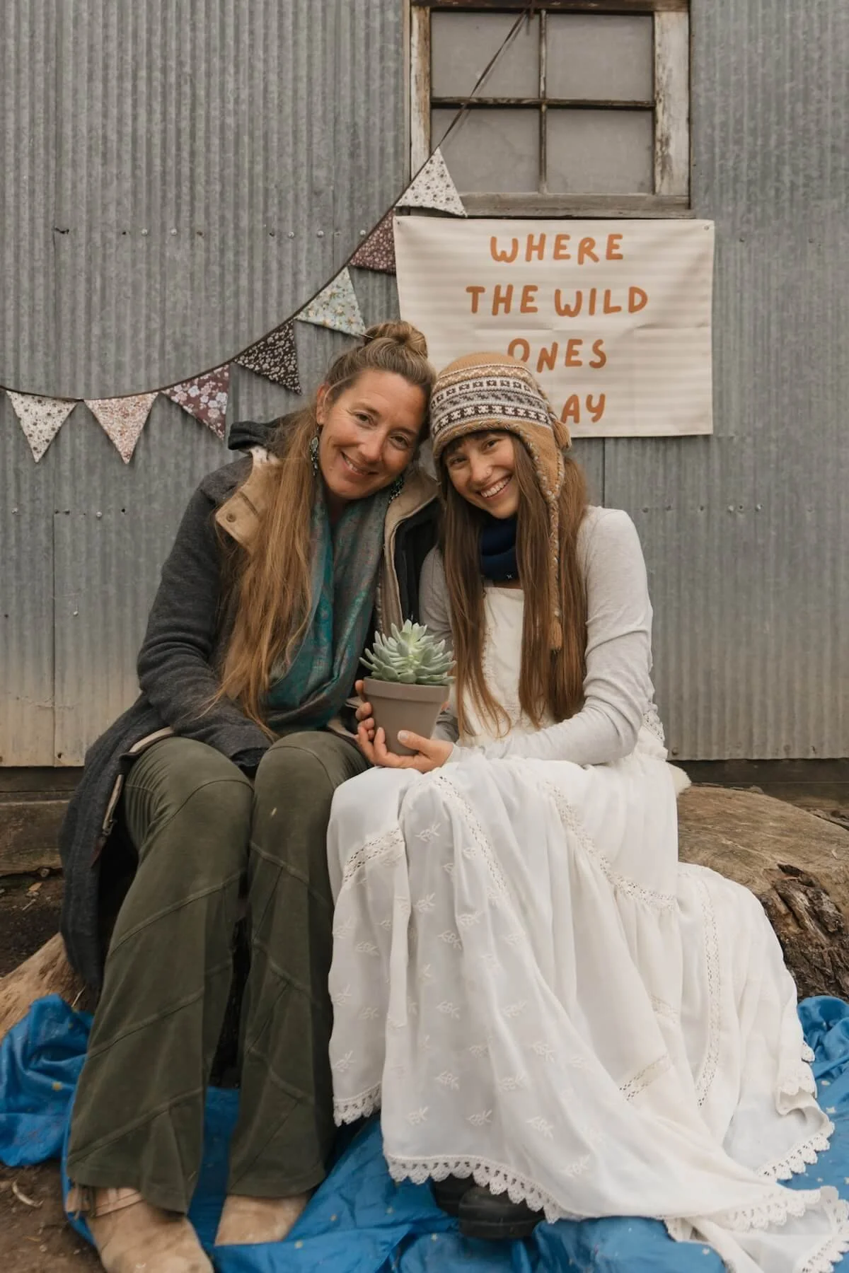 Two women sitting close together outdoors, holding a potted succulent plant. One woman is dressed in a white dress and wearing a knit hat, while the other is in casual clothing with a scarf. They are smiling in front of a corrugated metal wall with a sign that reads 'Where the Wild Ones Play' and a decorative bunting hanging above.