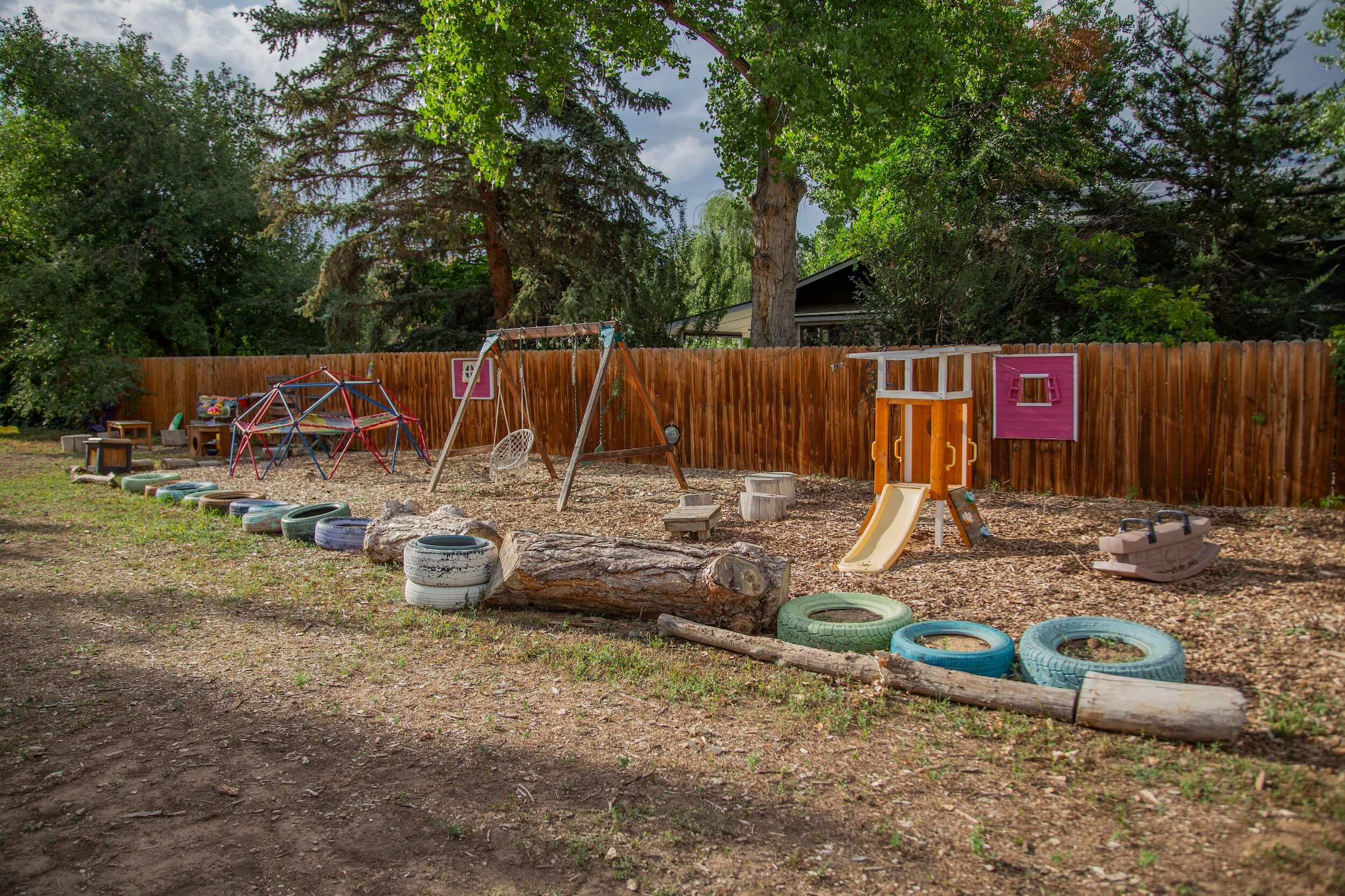 A backyard playground area with a wooden fence, trees, and play structures including swings, slide, and climbing frames. There are tires and logs on the ground.