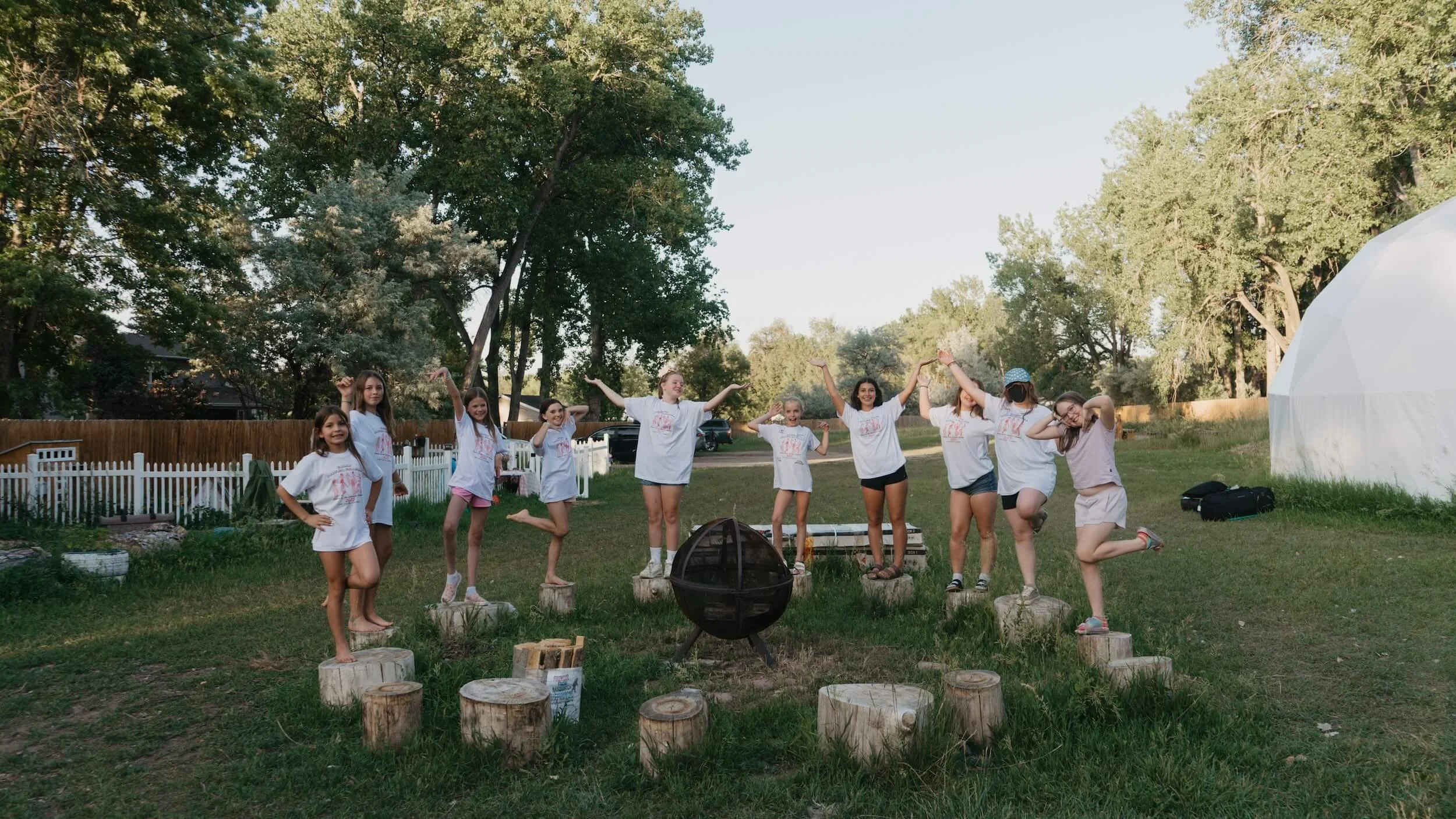 Group of young girls standing and posing outdoors in a backyard with trees, a white fence, and a white tent, enjoying a sunny day.