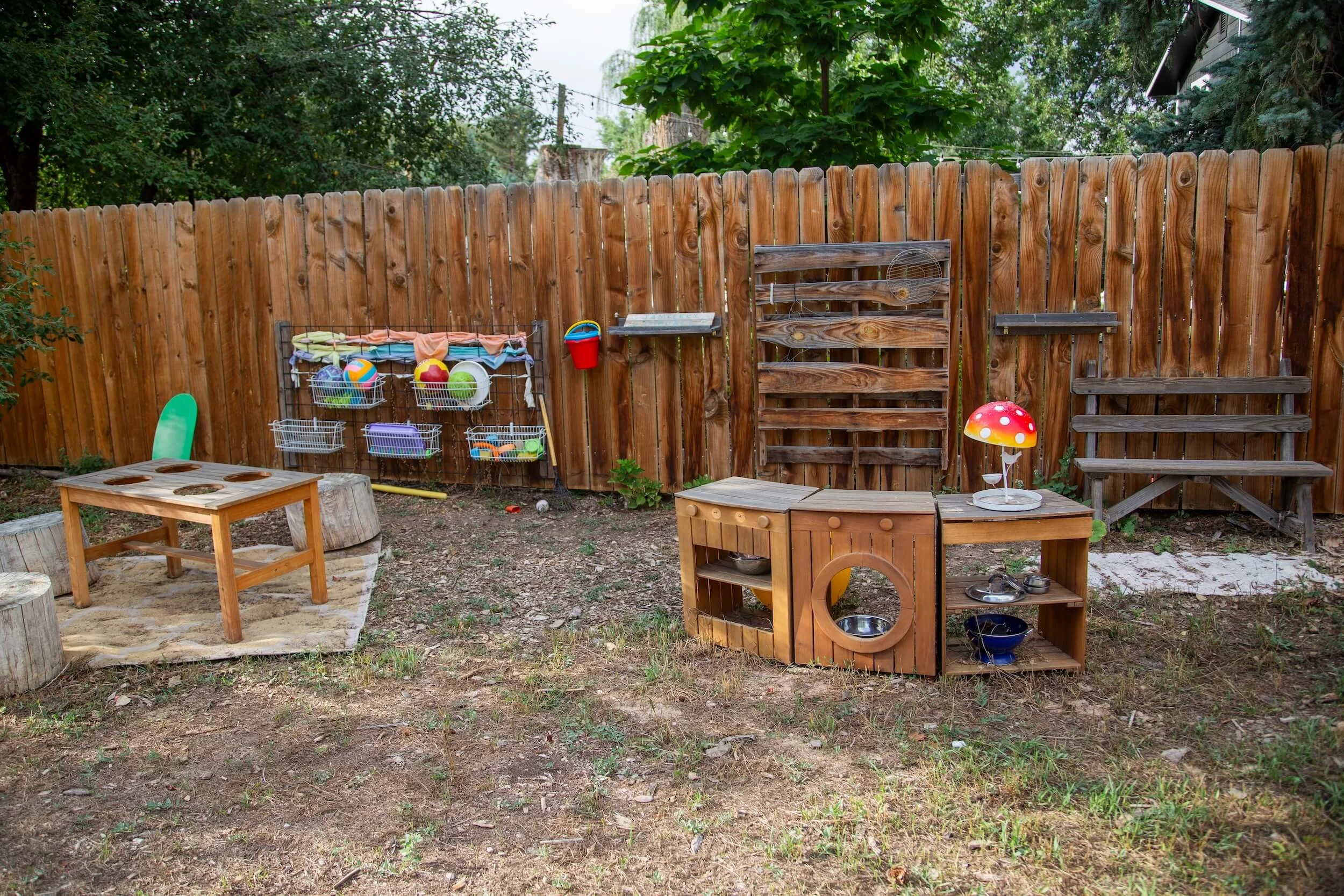 Backyard with a wooden fence, children's toys, tables, and a grassy, partially dirt ground surrounded by trees and greenery.