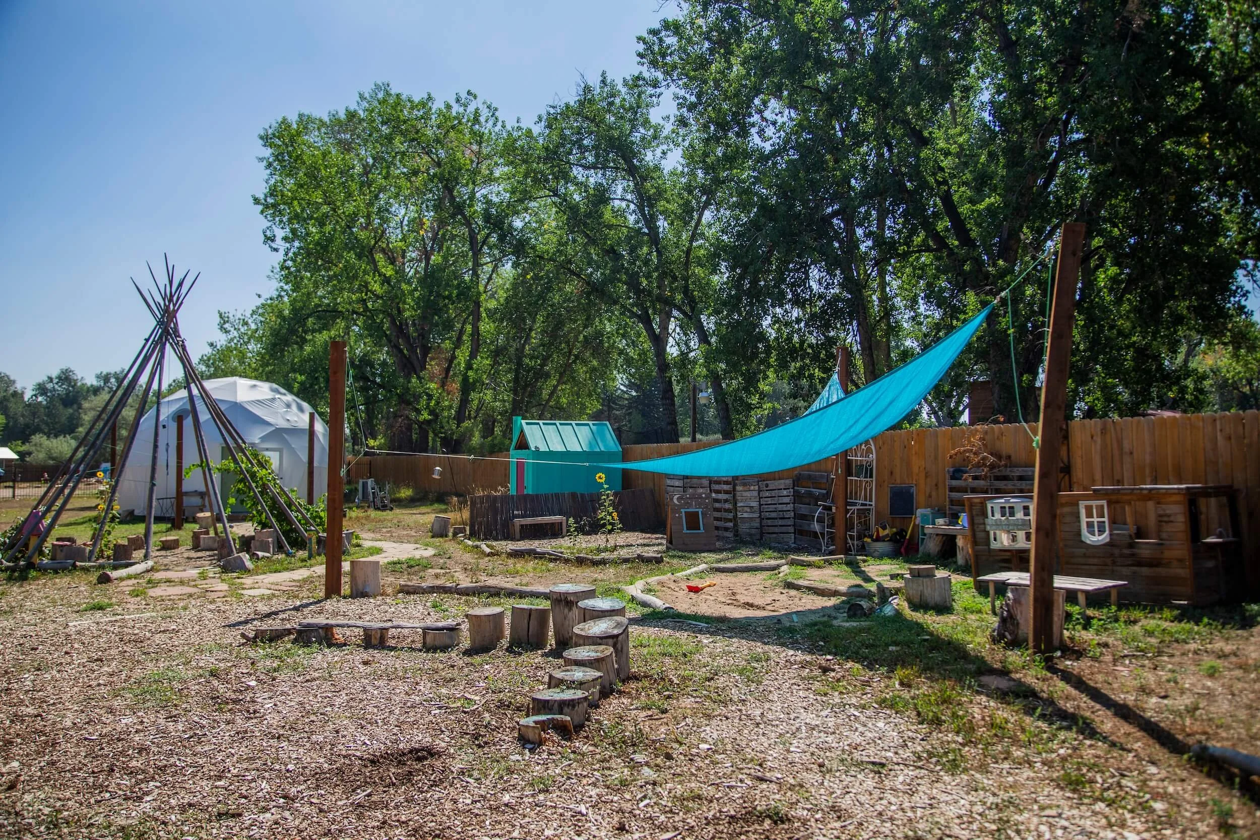 A backyard with a blue canopy, a small playhouse, a tipi made of wooden poles, a fence, and an outdoor space with trees and various objects scattered around.