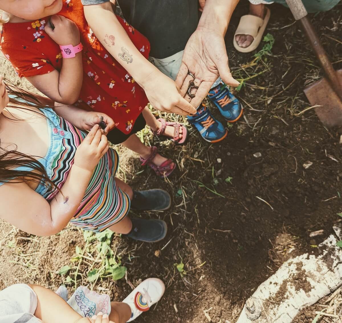 Children and an adult outdoors on soil. The adult is showing metal rings, children look on with curiosity. One girl holds a small object, all are wearing casual summer clothes and sandals.