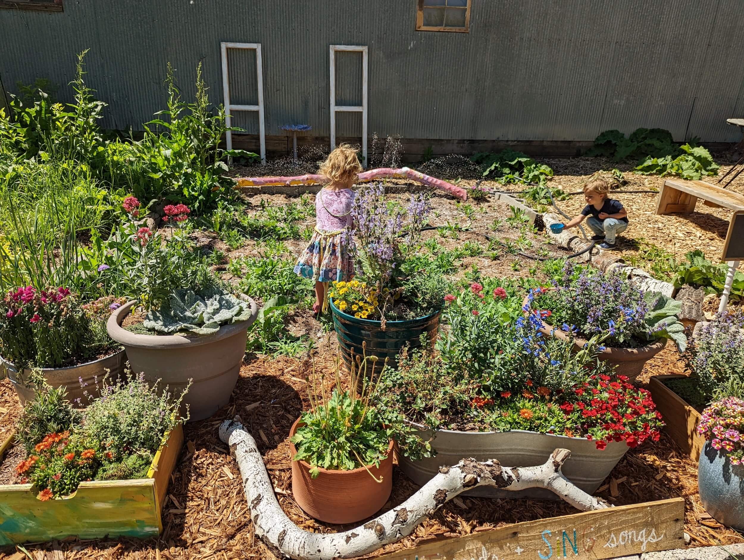 Two children in a garden with flowers, watering plants with hoses, surrounded by potted plants and garden decorations, against a house with corrugated metal siding.