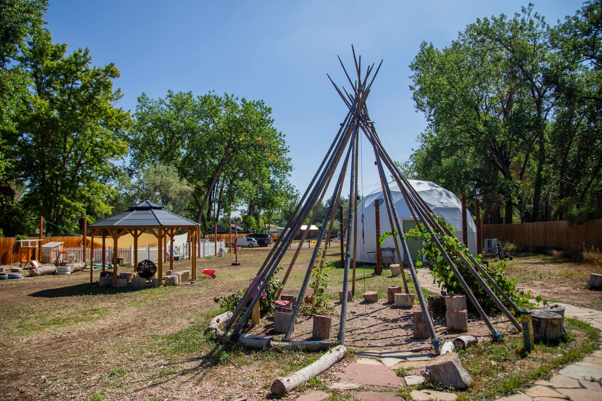 A backyard with a teepee structure made of wooden poles, a gazebo, a white dome-shaped tent, trees, and a wooden fence.