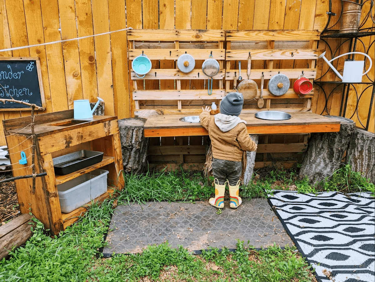 Child wearing rain boots and a beanie at a rustic outdoor kitchen station with hanging pots and pans, a sink, and gardening tools.