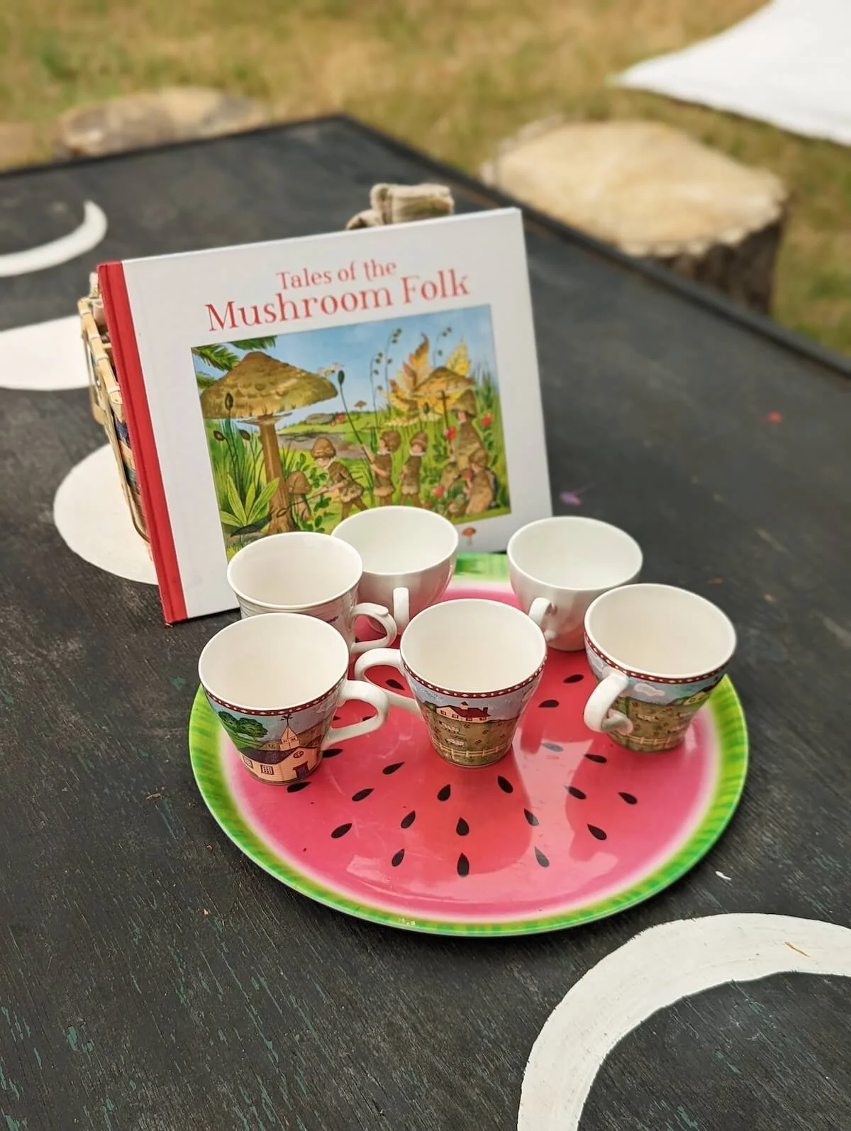 A novelty watermelon-shaped tray holding six ceramic cups with scenic village and countryside designs, set on a painted wooden table with a book titled "Tales of the Mushroom Folk" featuring an illustration of mushroom folk in a forest scene in the background.