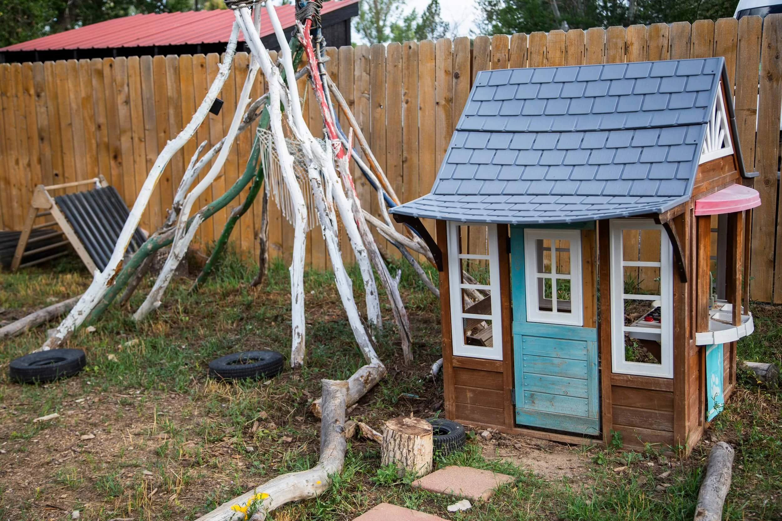 A backyard scene with a wooden fence, a small playhouse with a blue roof, and a pile of driftwood or stripped branches leaning against the fence.