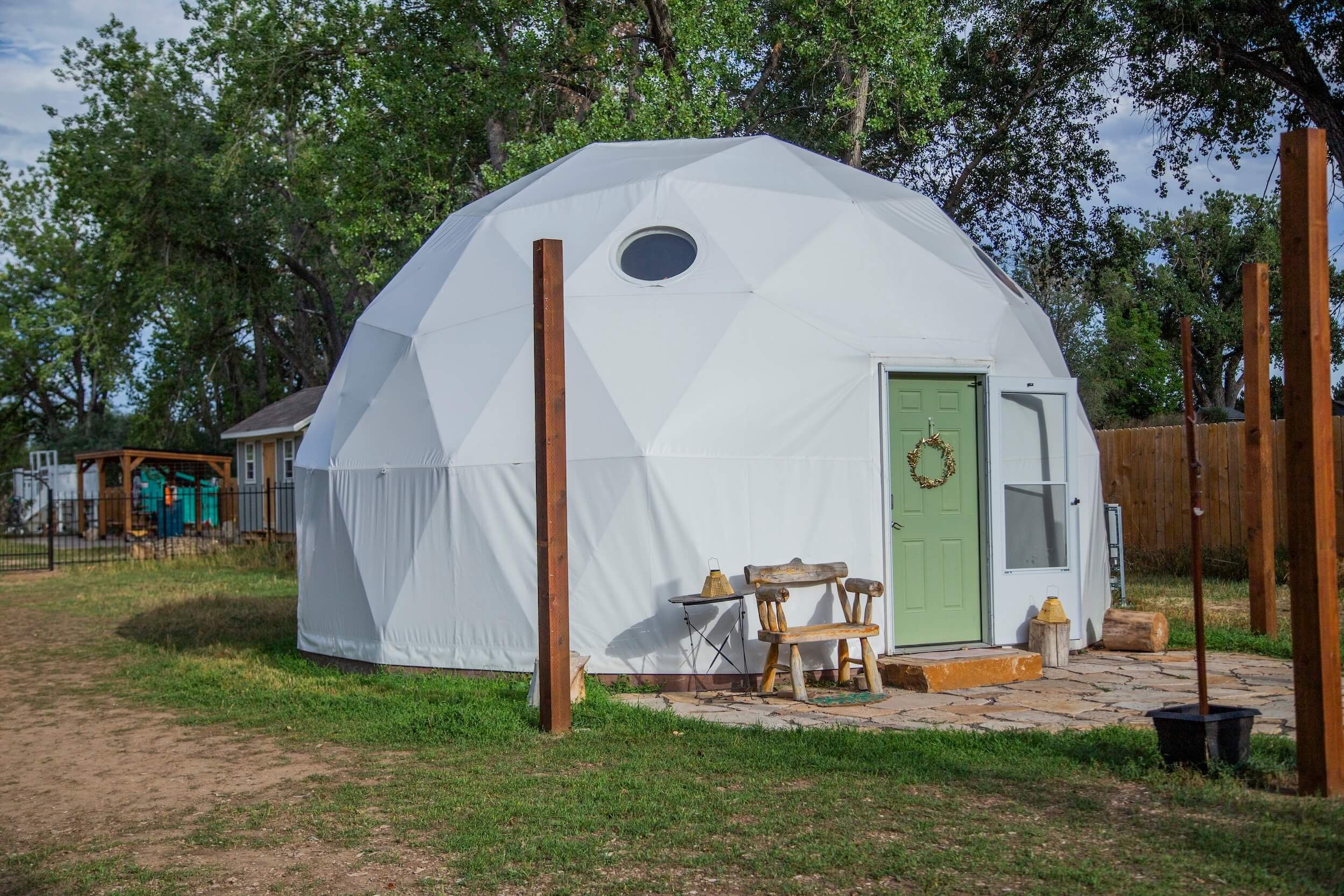 A white geodesic dome with a green door, a small window, and a wreath hanging on the door. The dome is set on a brick patio with outdoor furniture and a wooden fence surrounds the yard. There are trees and a cloudy sky in the background.