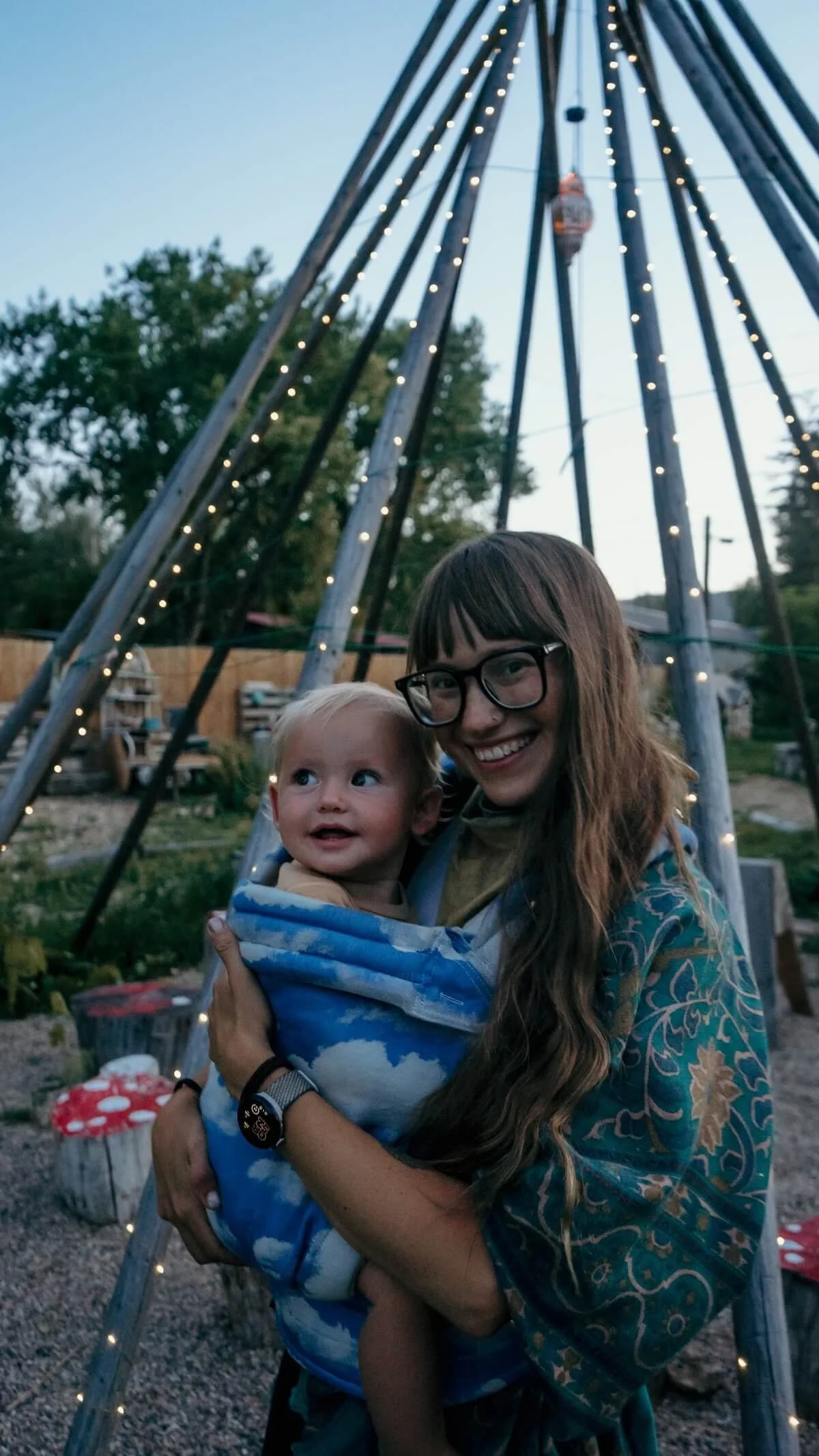 A woman with long hair and glasses holding a smiling toddler in a blue patterned baby carrier outdoors near a swing set decorated with string lights during evening.