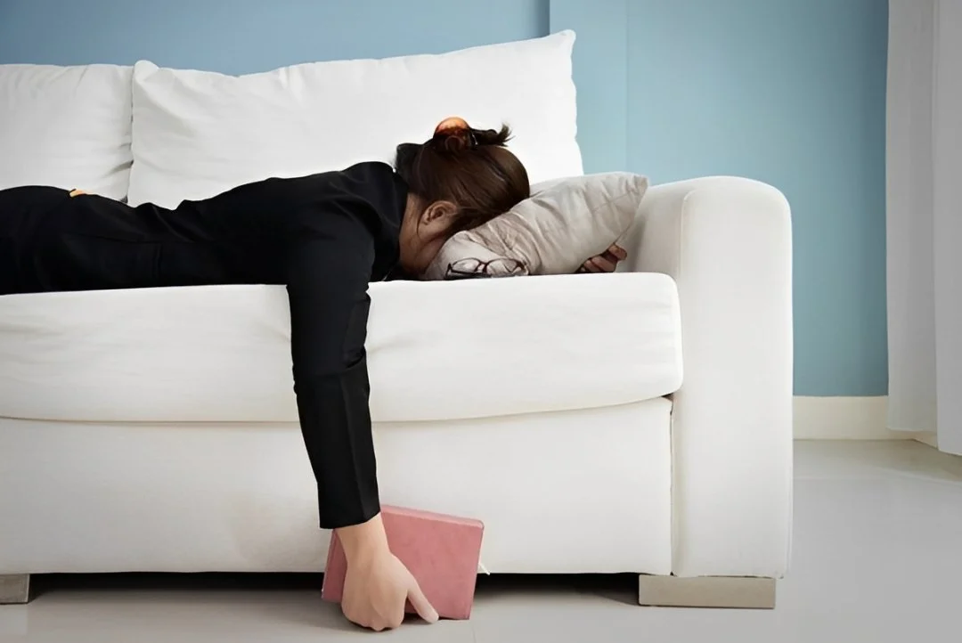 A woman with brown hair lying face down on a white couch with a beige pillow under her face, reaching down with one hand to pick up a pink book from the floor.