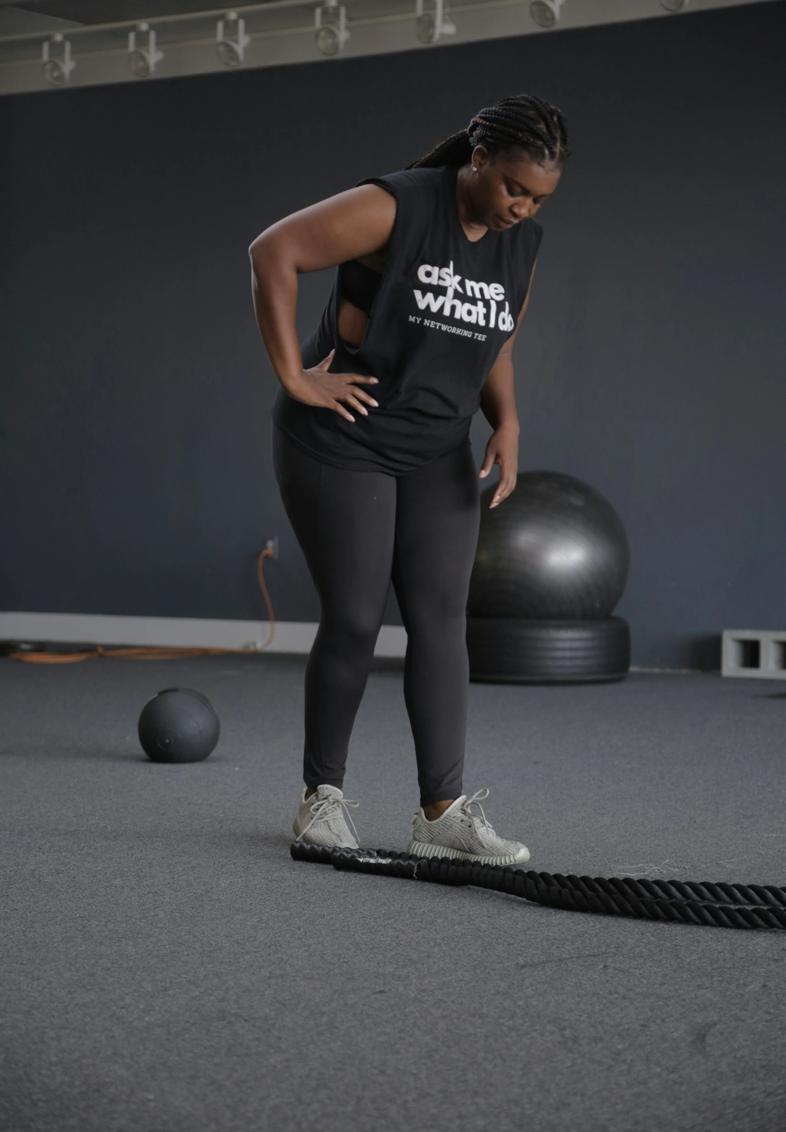 A woman in athletic clothing standing in a gym, looking down at a battle rope on the floor, with a large exercise ball and small medicine ball in the background.