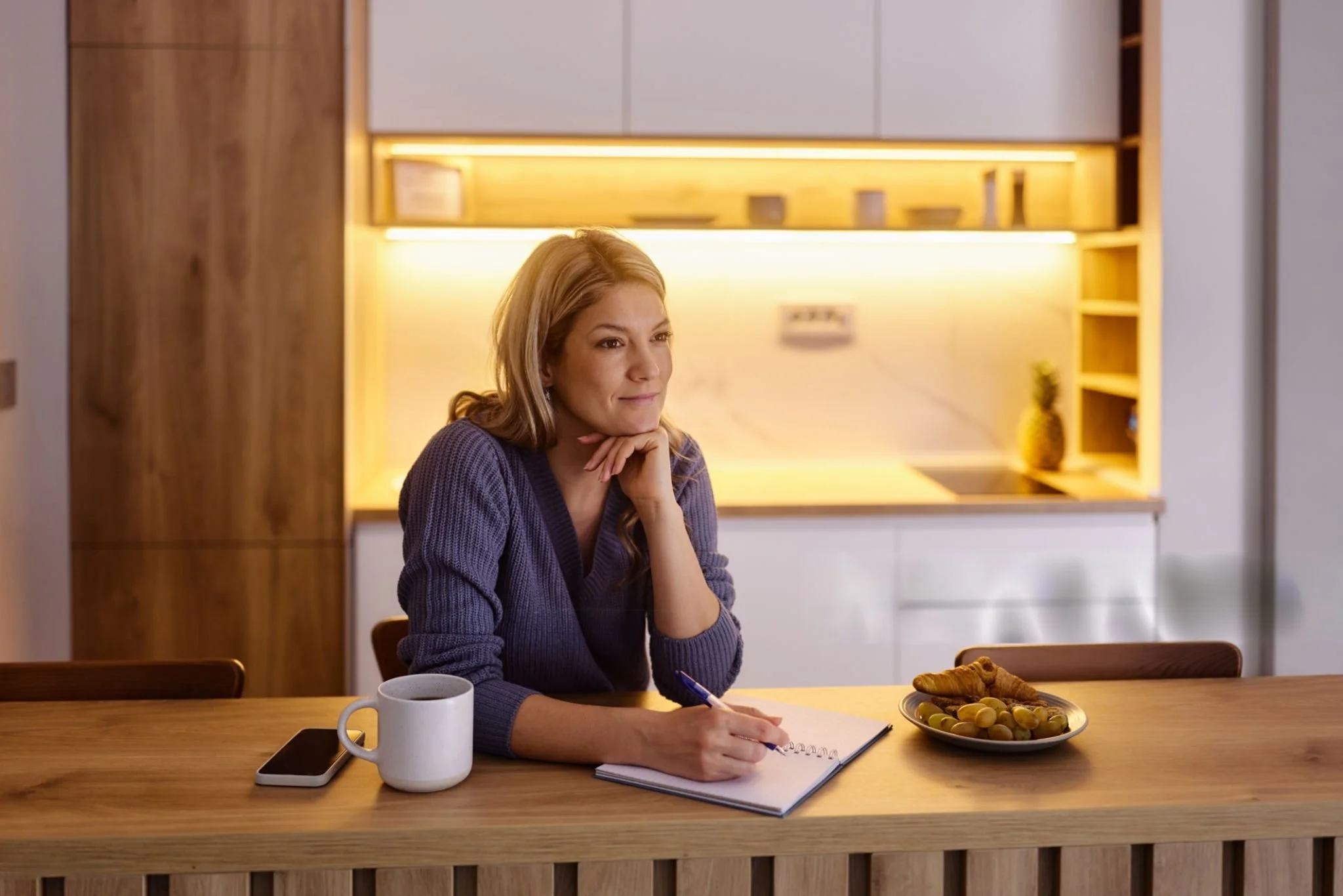 A woman with long, wavy blonde hair, wearing a purple sweater, sits at a wooden kitchen counter, holding a pen and writing in a notebook. On the counter in front of her are a white mug, a smartphone, and a plate of croissants and grapes. She appears to be in a modern, well-lit kitchen with wooden and white cabinets, and a pineapple on the counter in the background.