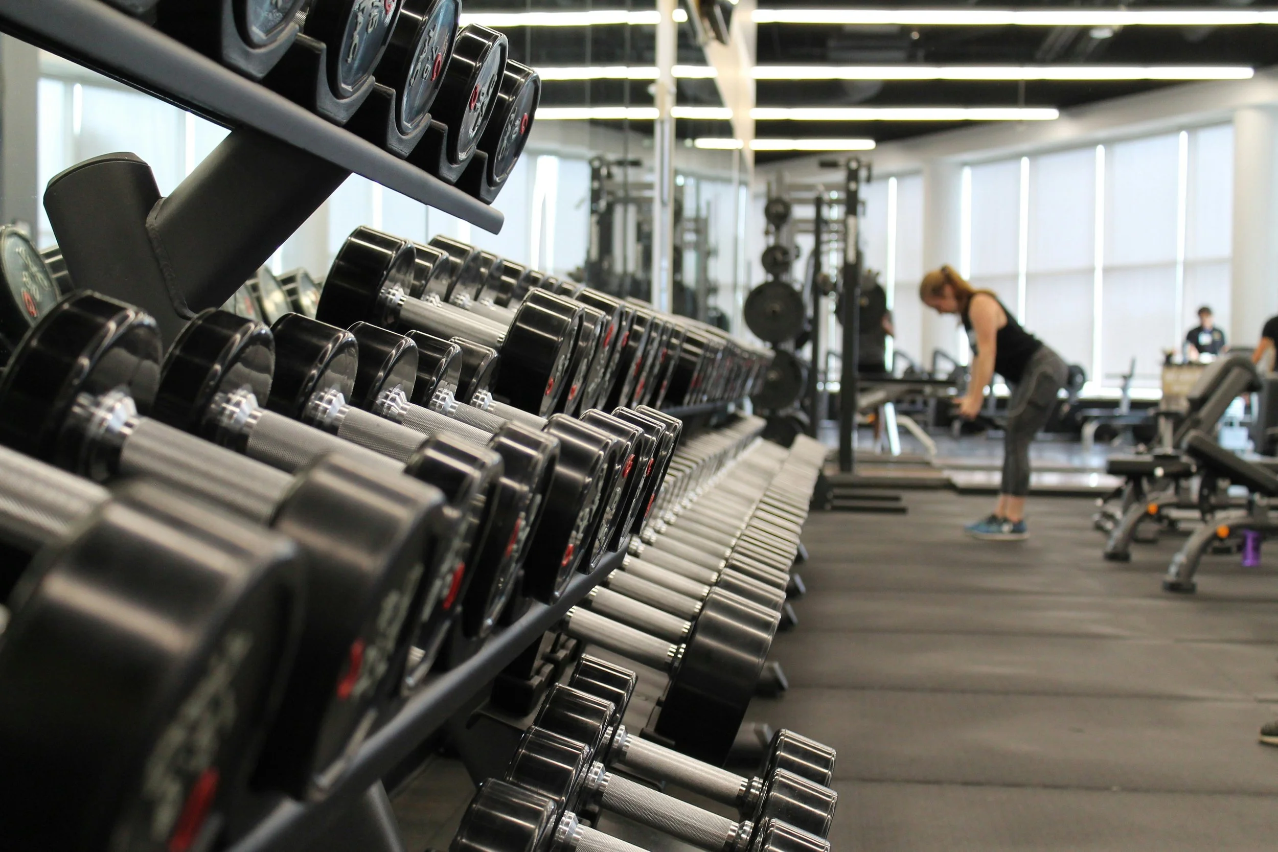 Row of dumbbells in a gym with a woman working out in the background.
