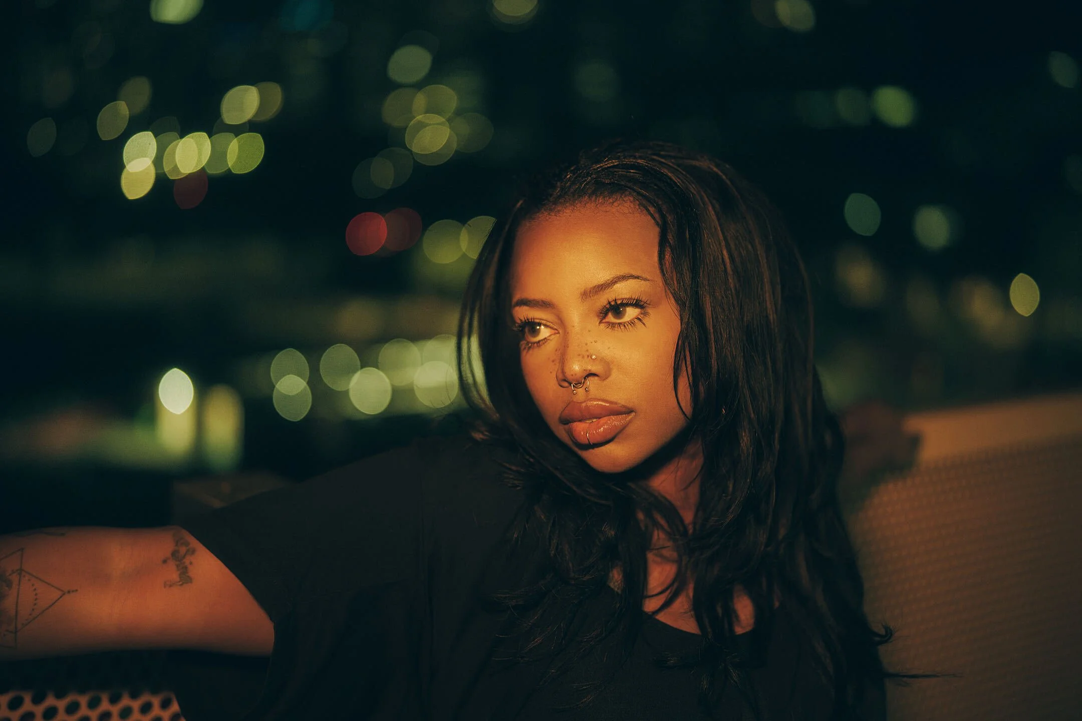 A woman with dark hair and freckles, wearing makeup and a septum piercing, gazing to her right, with blurred lights in the background.