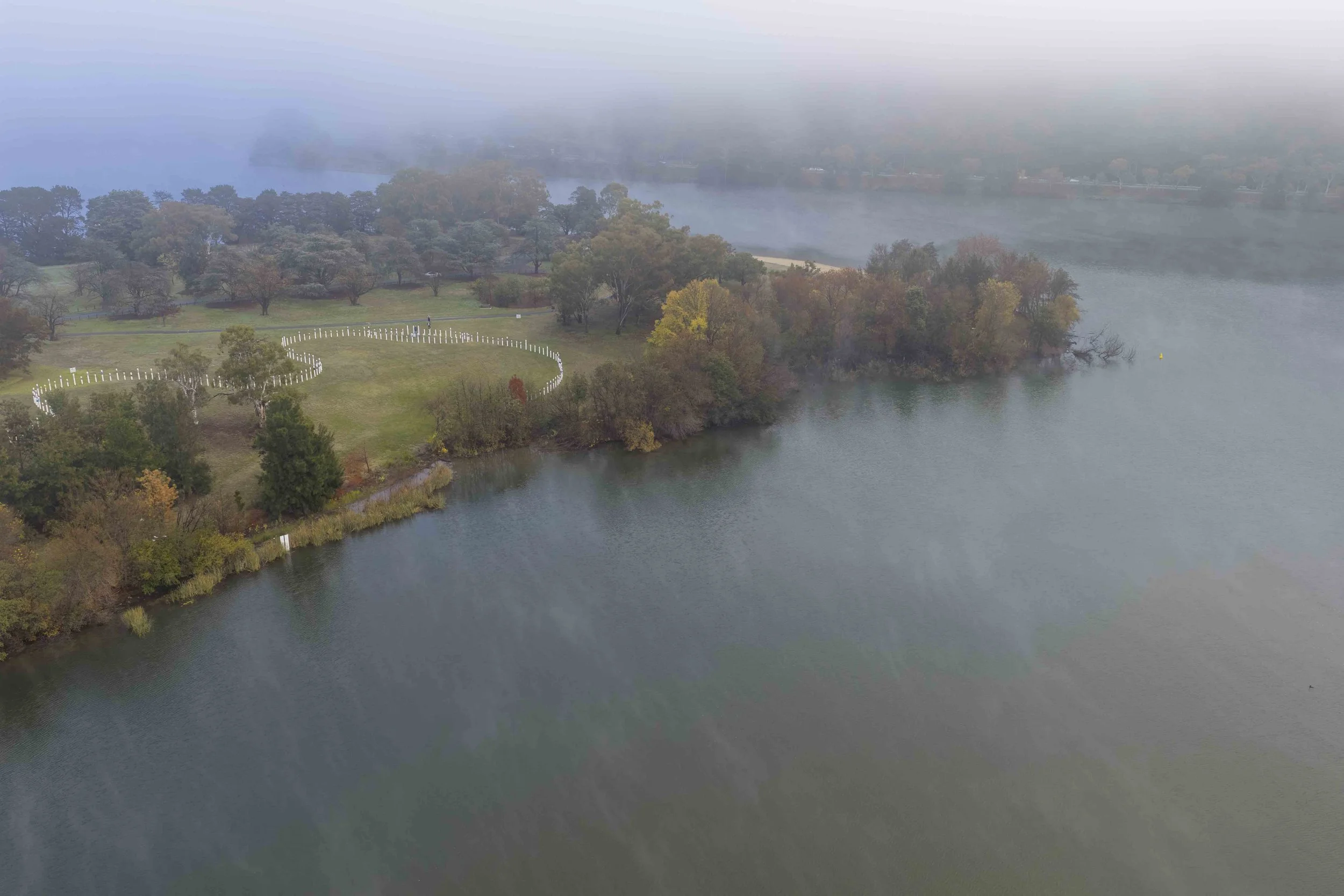 A foggy, lush green park with a circular pathway and white fence surrounding the grassy area, adjacent to a large body of water.