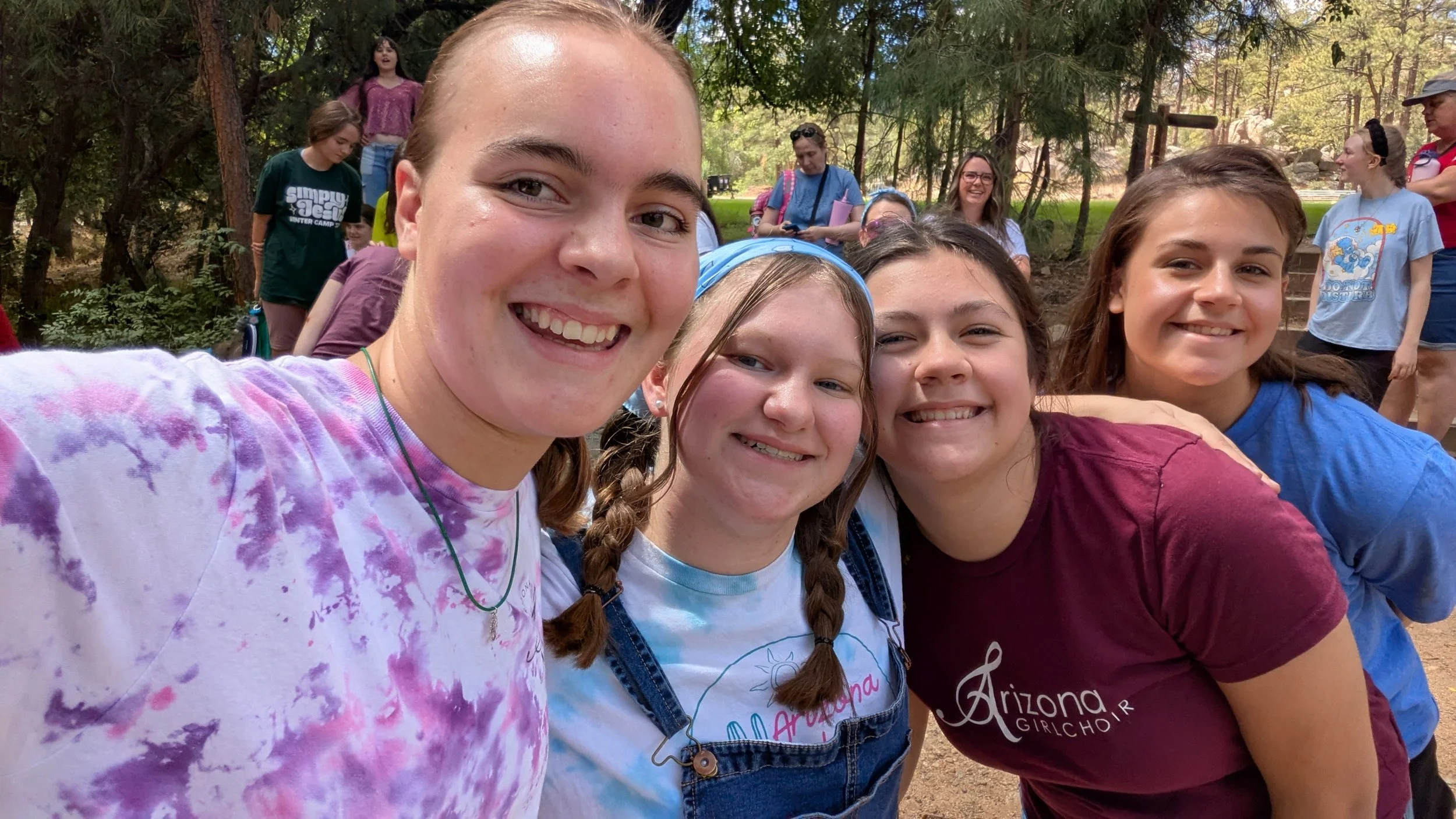A group of young girls smiling for a close-up selfie outdoors among trees, with other girls in the background.