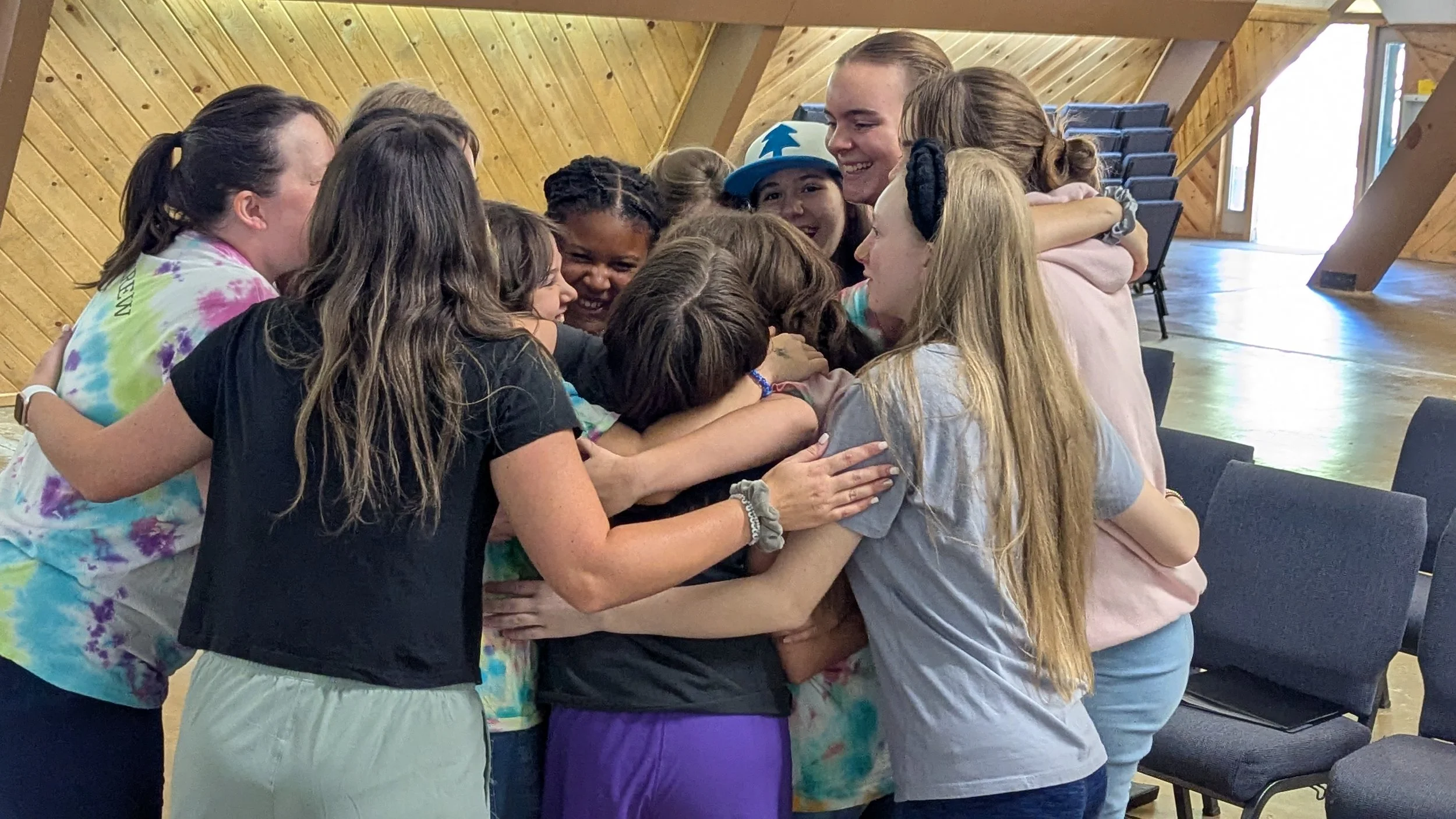 A diverse group of children and teenagers hugging each other in a close circle inside a wooden hall, smiling and showing affection.