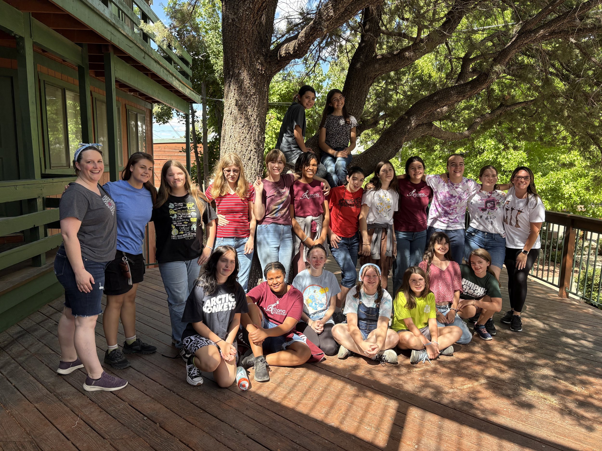 A group of children and teenagers gathered outdoors under a large tree, smiling and posing for a picture on a wooden deck. The background features a building with green frames and windows, and the weather is sunny with a blue sky.