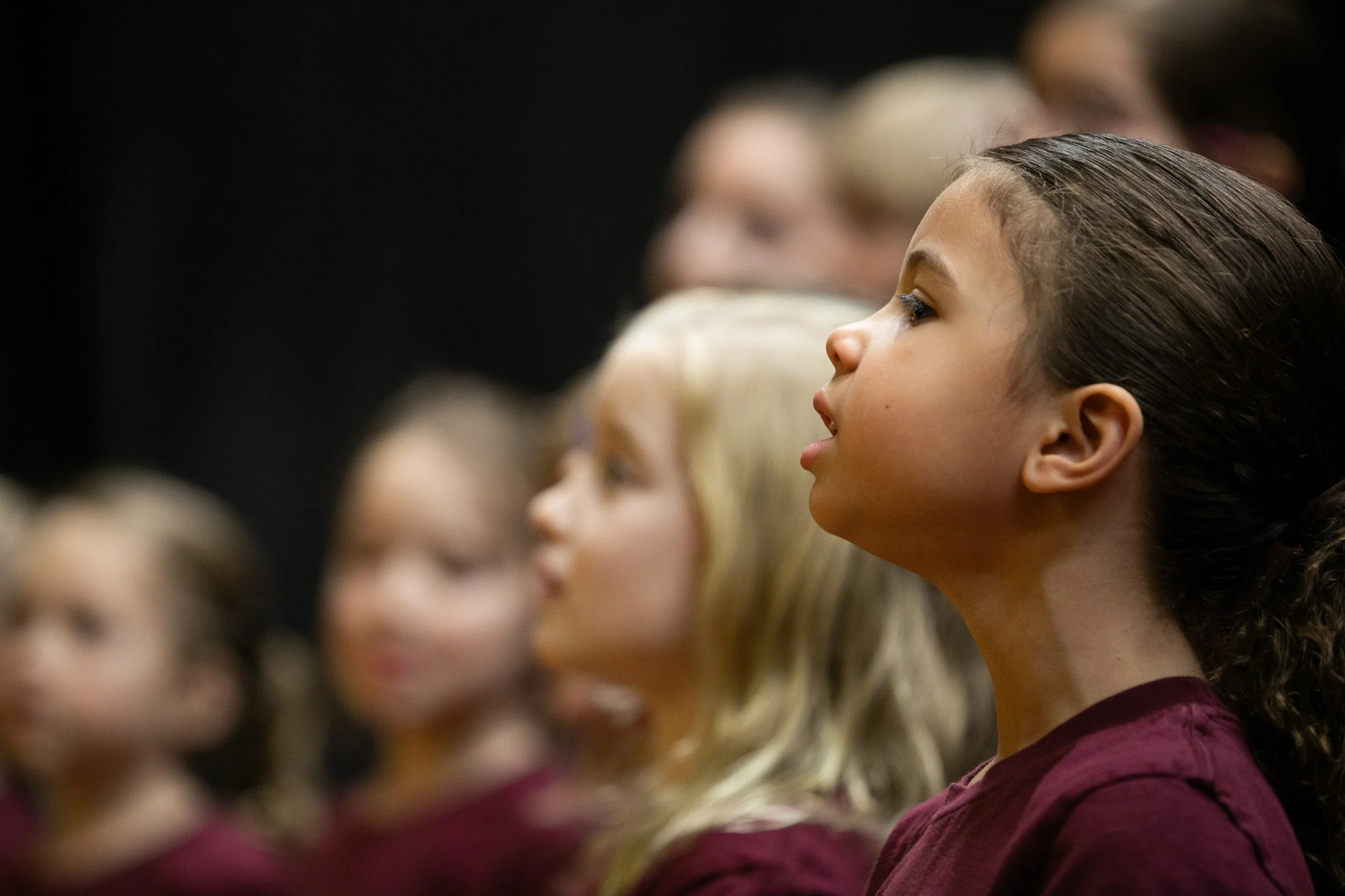 Young girl with dark, wet hair and a maroon top, sitting in a row of children at an event, listening attentively.