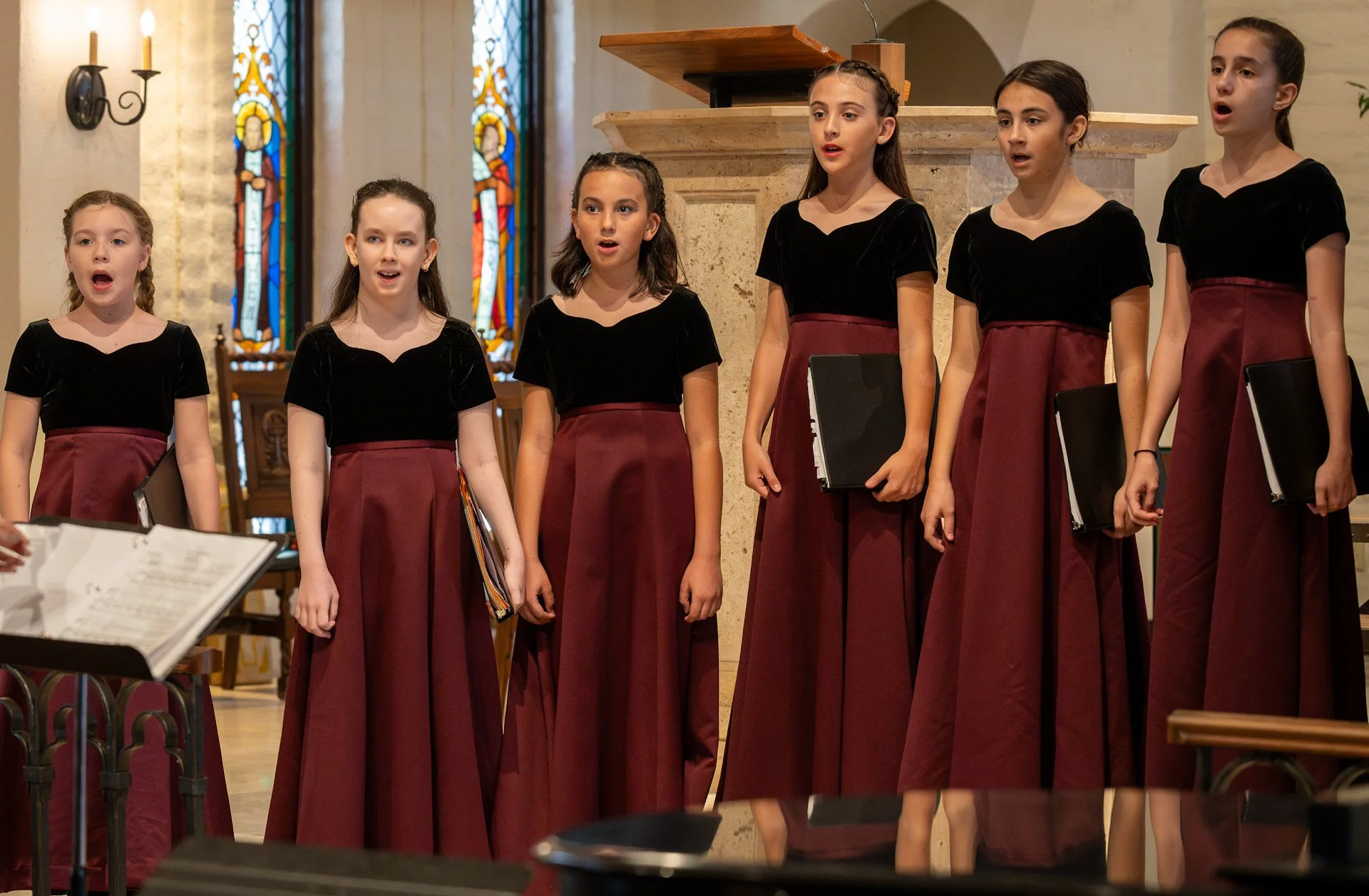 Six young girls singing in a choir inside a church, wearing black tops and long maroon skirts, with stained glass windows in the background.