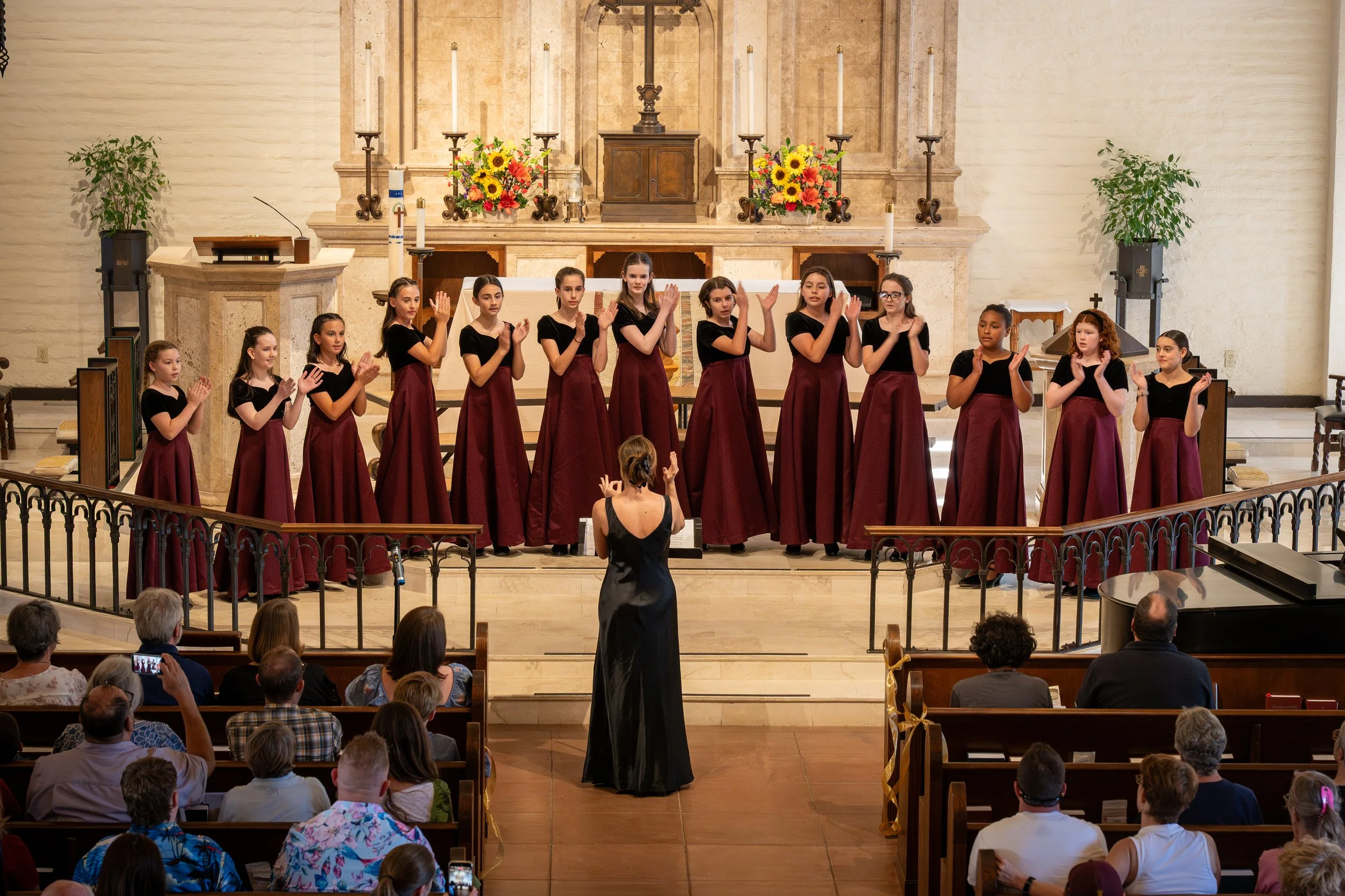 A choir of young girls dressed in black shirts and maroon skirts sing on a church altar. The choir is led by a conductor in a black dress, standing in front of the group. The church interior features a large altar decorated with flowers and candles, with two tall potted plants on either side. Several people in the audience are watching and some are taking photos.