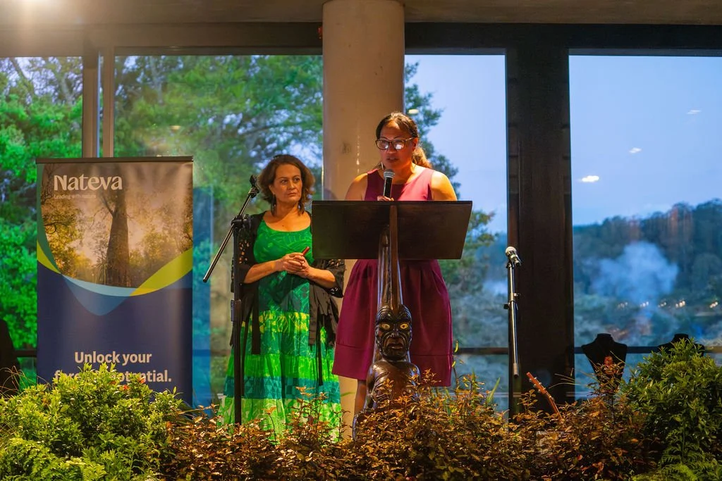 Ramona Radford awards Shontelle Bishara the Resilience award at Ngahere Māori Awards (Māori forestry). 'Nateva' banner with 'Unlock your potential', in background, decorative plants in front. Large windows show trees and a partly cloudy sky outside.