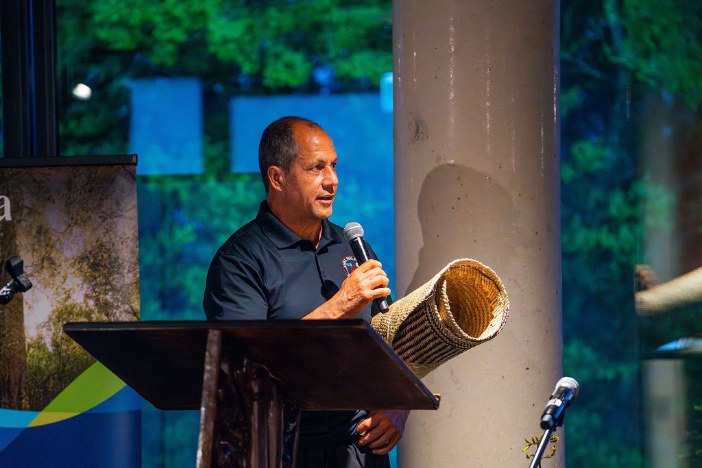 Doug Macredie receiving his award holding his award which is a weaved mat, or whāriki, for Transition Forestry during Ngahere Māori Awards 2025, at a podium, with a large column and greenery in the background. Doug was also awarded $10,000.