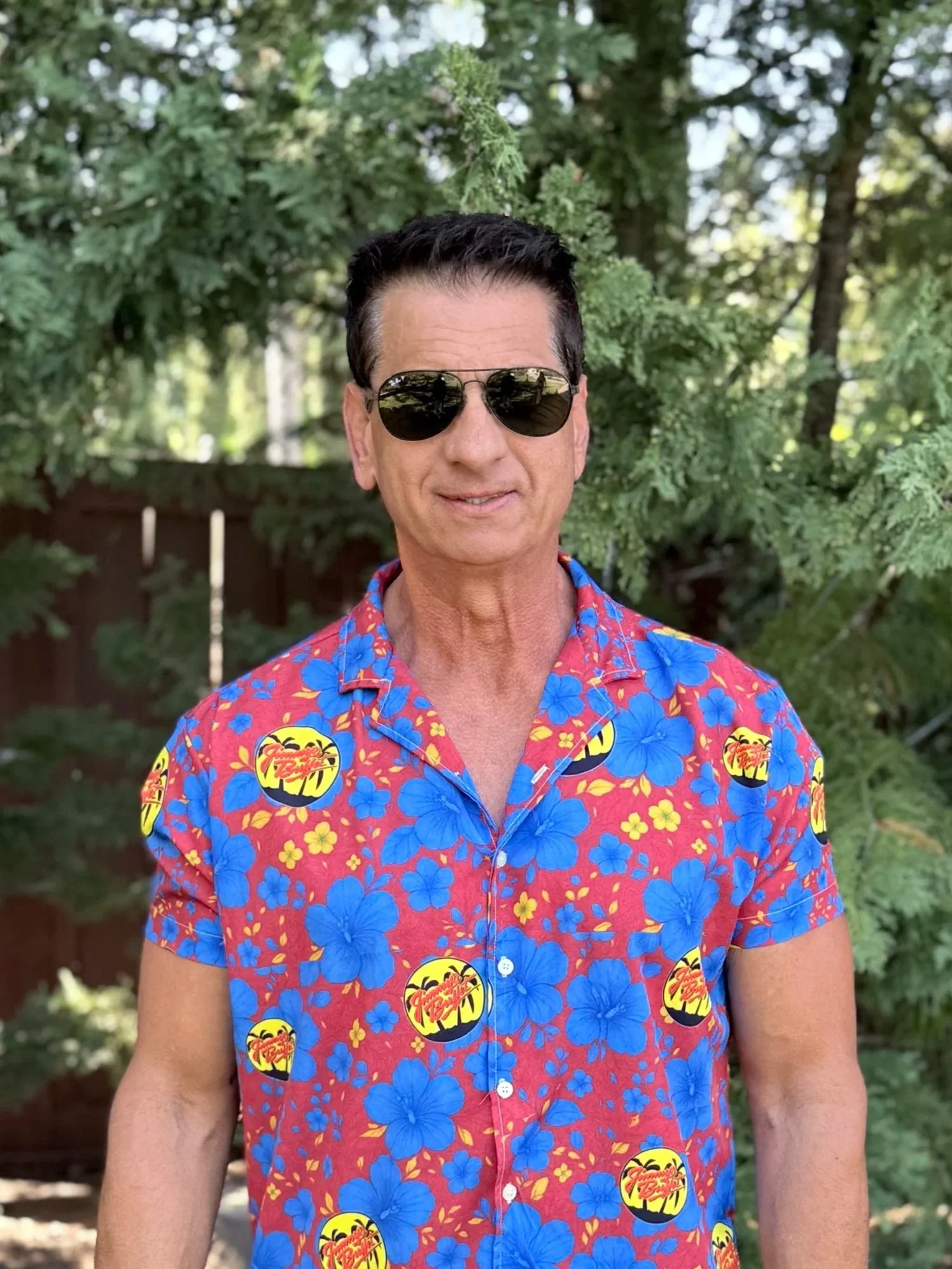 Man wearing a white hat with a red and yellow logo, black sunglasses, a blue jacket, and a beaded necklace, standing in front of a neon sign and wooden wall.