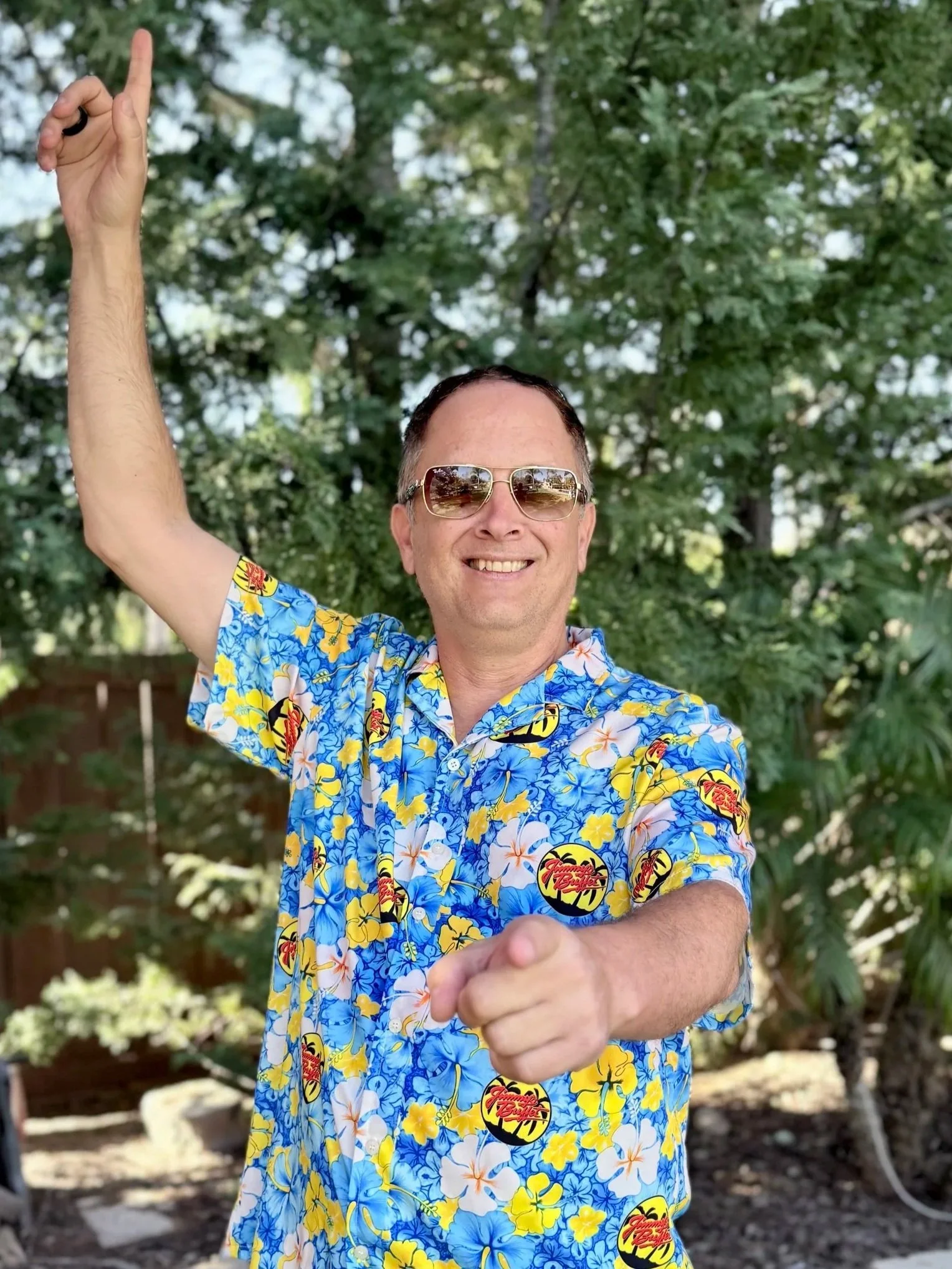 A smiling man wearing a black baseball cap and a blue and white jacket with a red logo, standing in front of a colorful tropical-themed background.