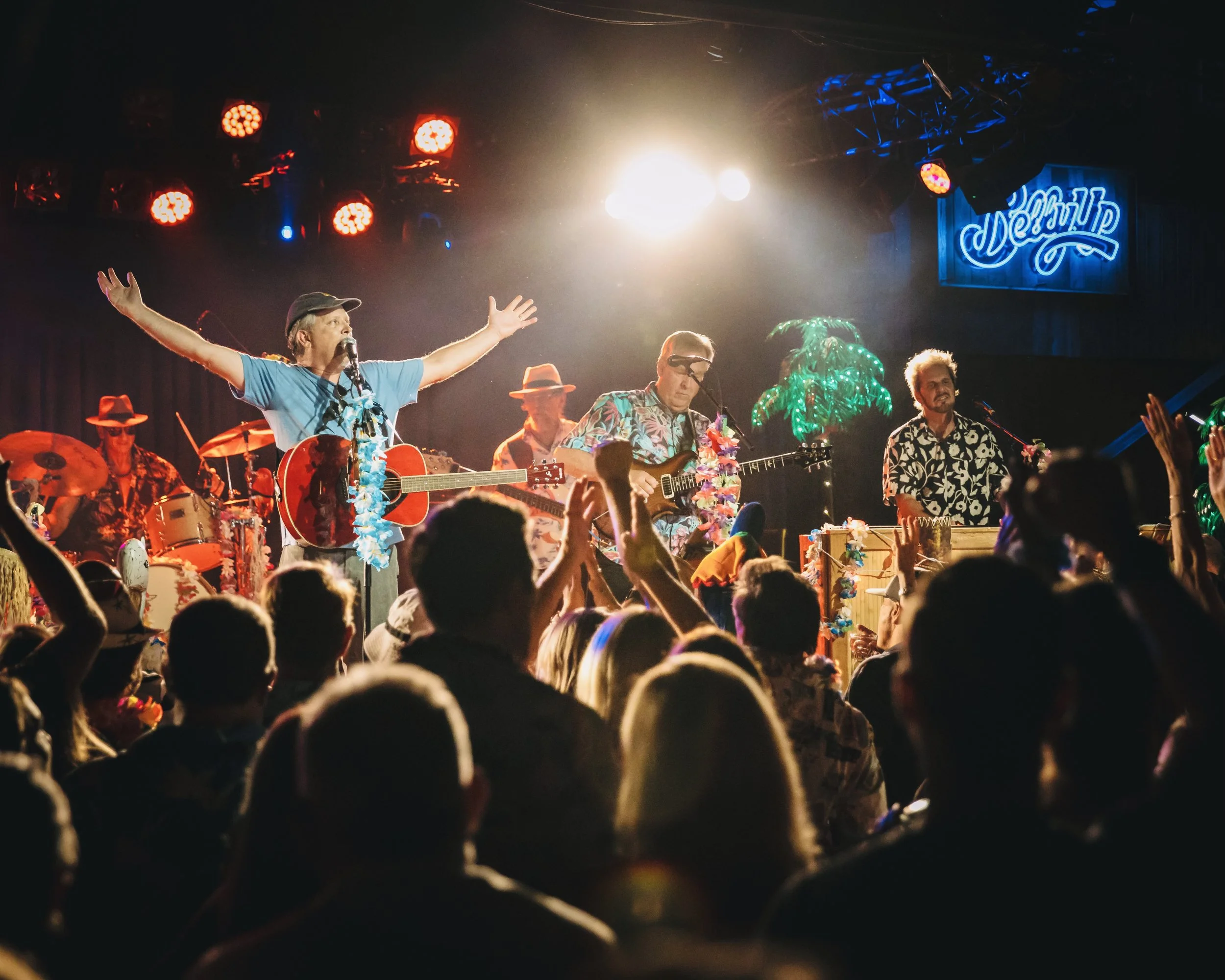 A live concert on a tropical-themed stage with band members playing guitars, drums, and percussion, audience with raised hands, stage illuminated with red and white lights, sign reading 'Deep Dive' in the background.