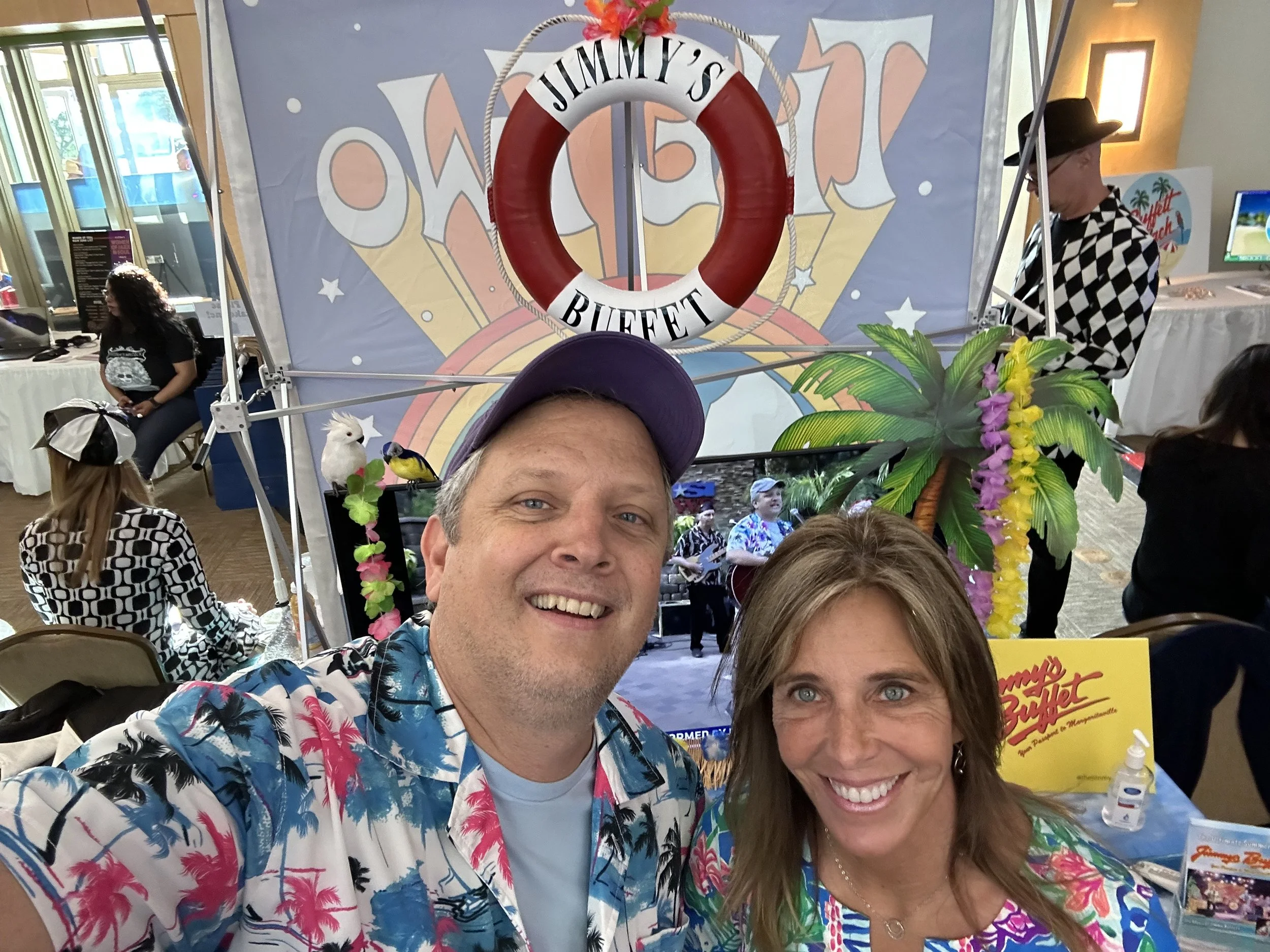 A smiling man and woman dressed in tropical shirts taking a selfie at a Jimmy Buffett-themed event. The background features a stage with performers, tropical decorations, a large flotation device with "Jimmy's Buffett" written on it, and other attendees enjoying the event.
