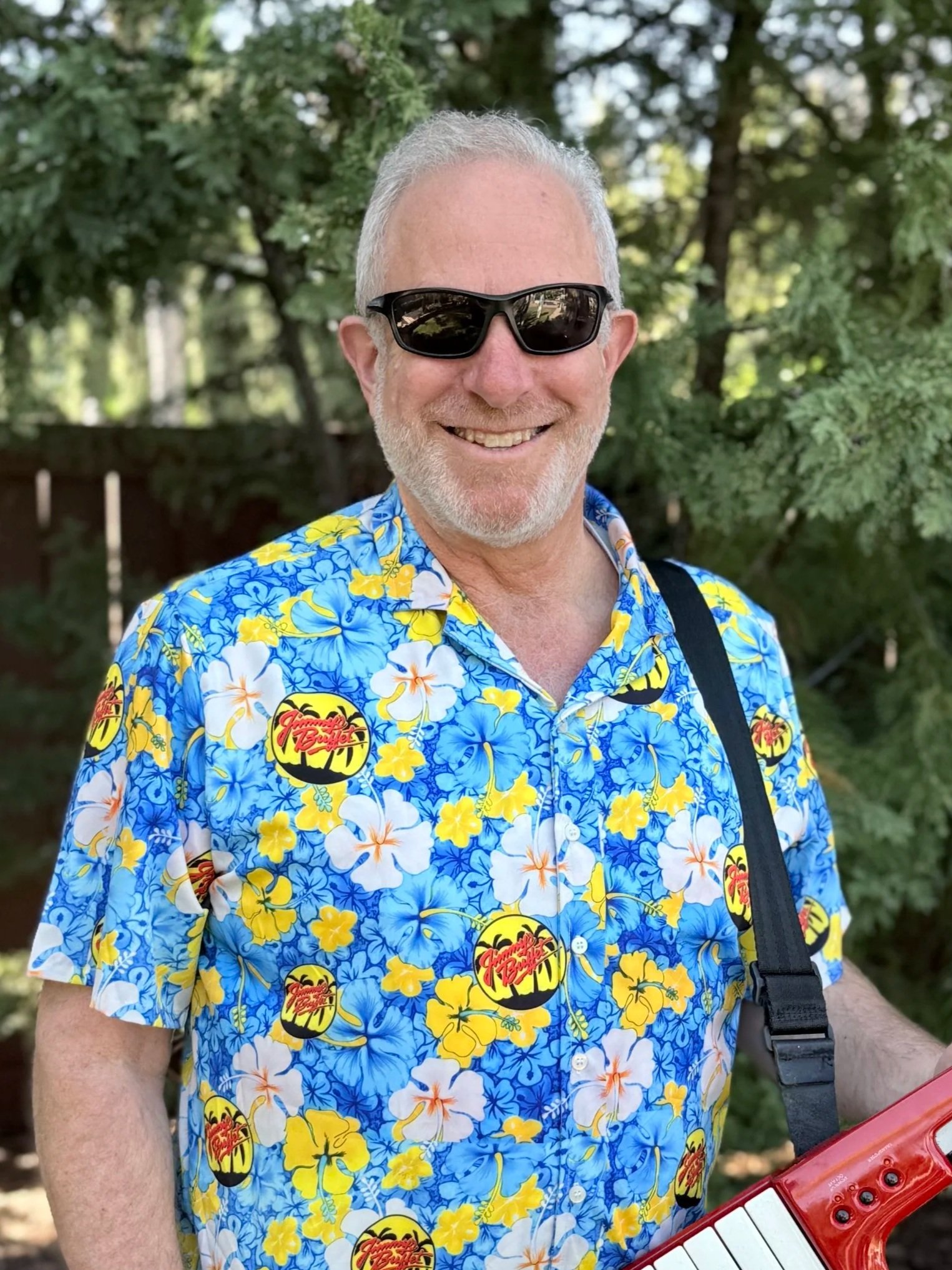 Smiling man with long curly hair, sunglasses, and a blue and white jacket, pointing at the camera outdoors.