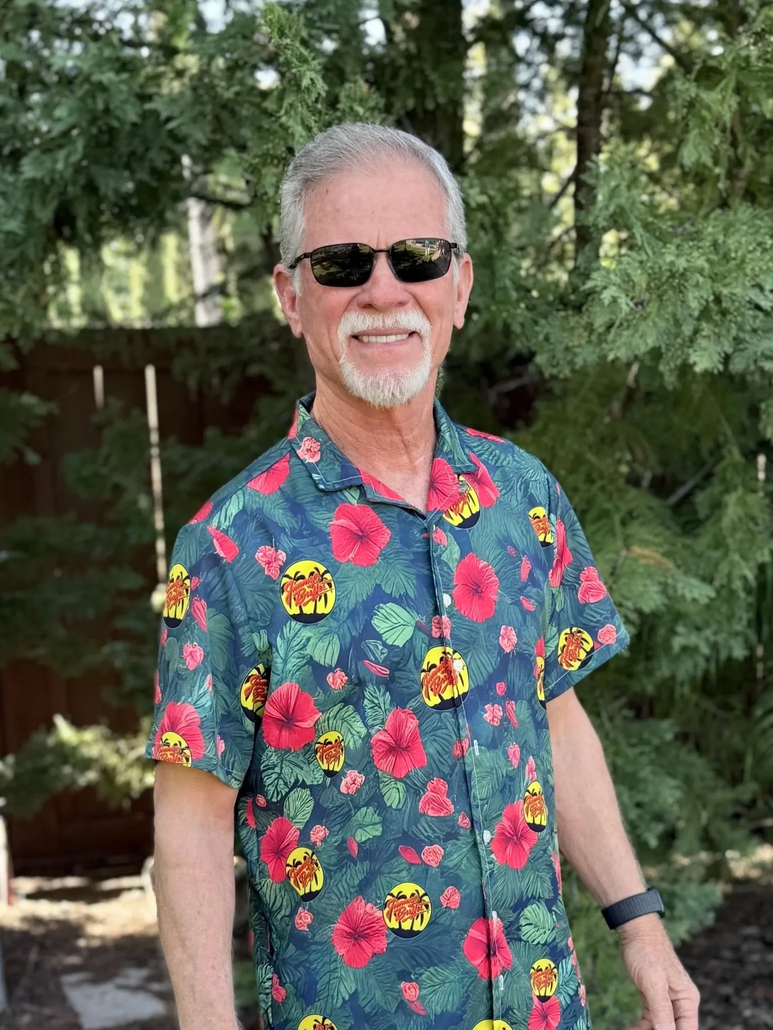 Close-up of an older man with gray hair and a beard, smiling, wearing a blue and white tie-dye jacket with red lettering, standing outdoors near trees and a Christmas tree.