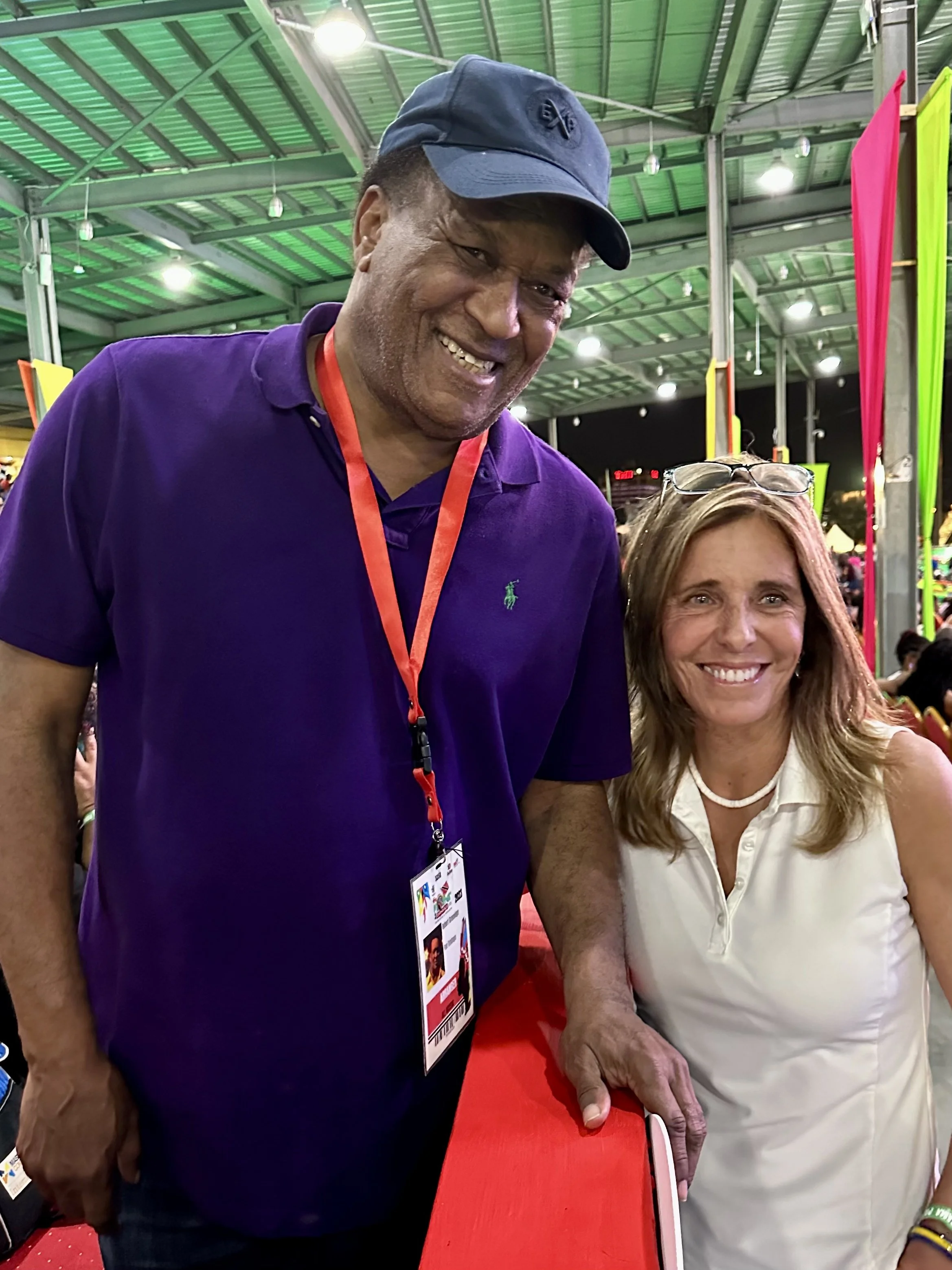 A man and a woman smiling at an outdoor event during the evening. The man is wearing a navy cap, purple polo shirt with a green logo, and has a conference badge around his neck. The woman is wearing a white sleeveless top, has sunglasses resting on her head, and a necklace. They are standing under a green metal roof with colorful banners in the background.