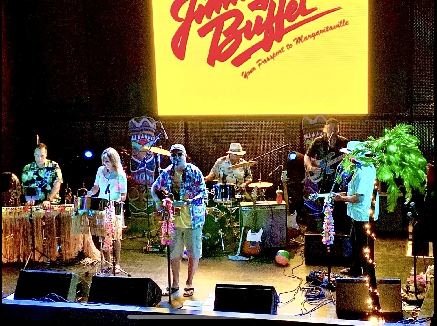 A band performing on stage with tropical and summer-themed decorations. Musicians are playing instruments such as drums, guitar, and keyboard, with a large yellow screen behind them displaying red text that says 'Jimmy Buffett' and what appears to be a tagline. The stage has Hawaiian leis, a small palm tree, and tiki totem decorations suggesting a tropical, beach party vibe.