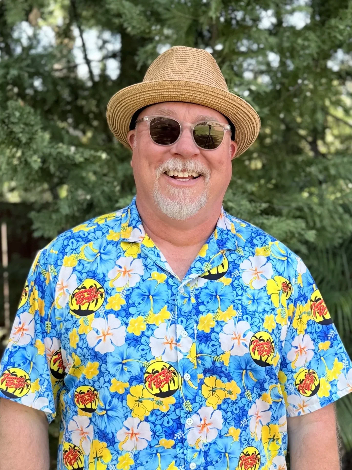 A man with a beard and glasses wearing a white cap and a blue colorful shirt, outdoors with greenery in the background.