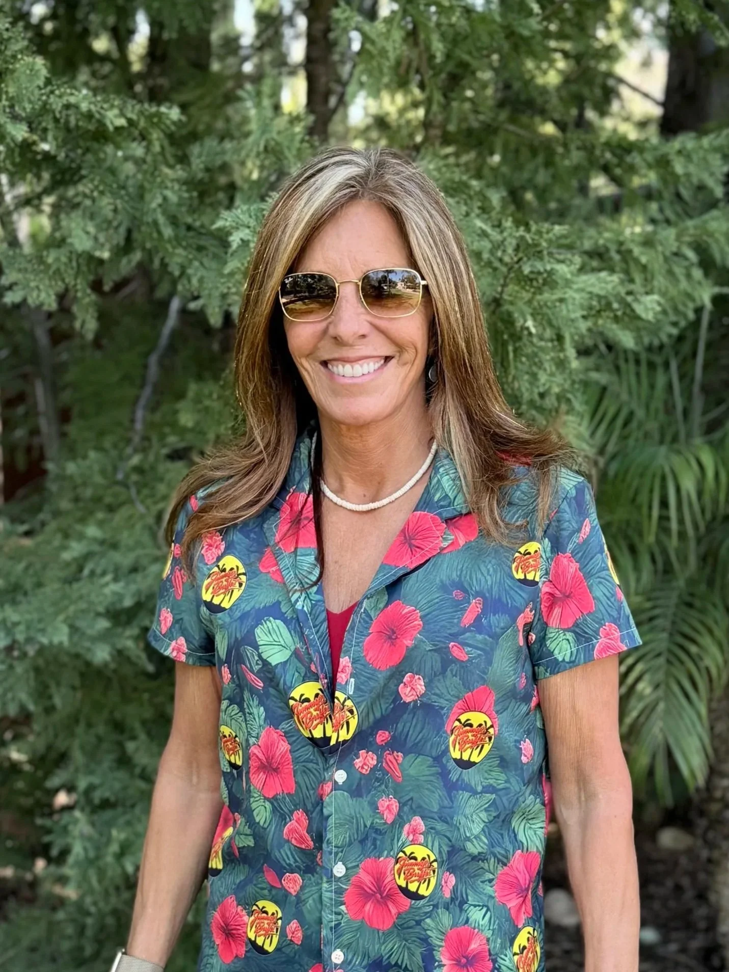 A woman smiling, wearing sunglasses, a black long-sleeve shirt, and a colorful tropical print shirt with palm trees. She has earrings and a star-shaped necklace. The background shows a sign that reads 'KUSH NEWS' and outdoor decorations.