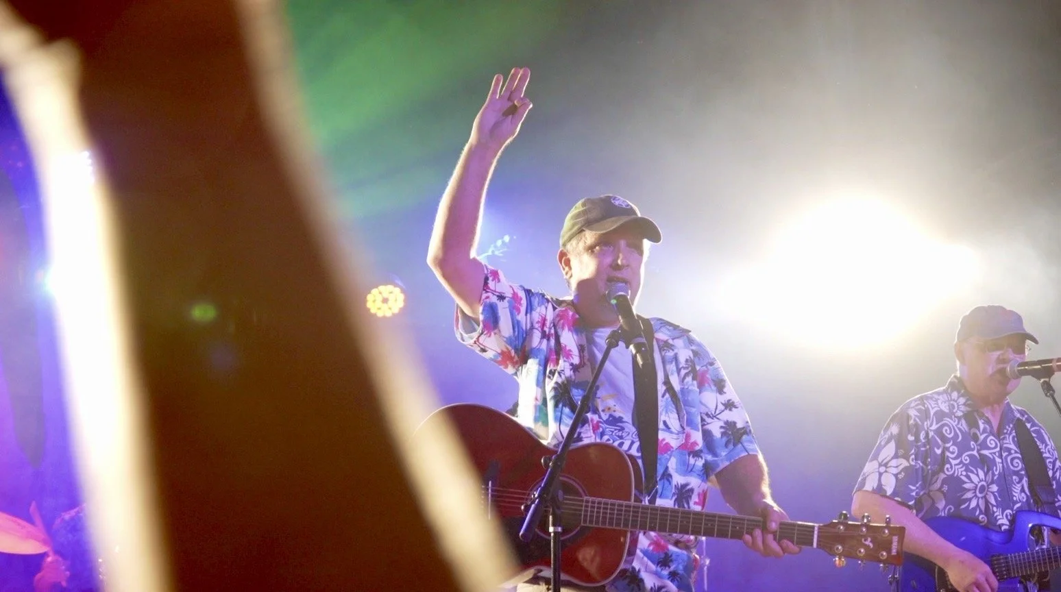 Two musicians performing on stage, one wearing a colorful floral shirt and a hat, playing an acoustic guitar, and the other in a patterned shirt and glasses, singing into a microphone. Bright stage lights in the background create a vibrant atmosphere.