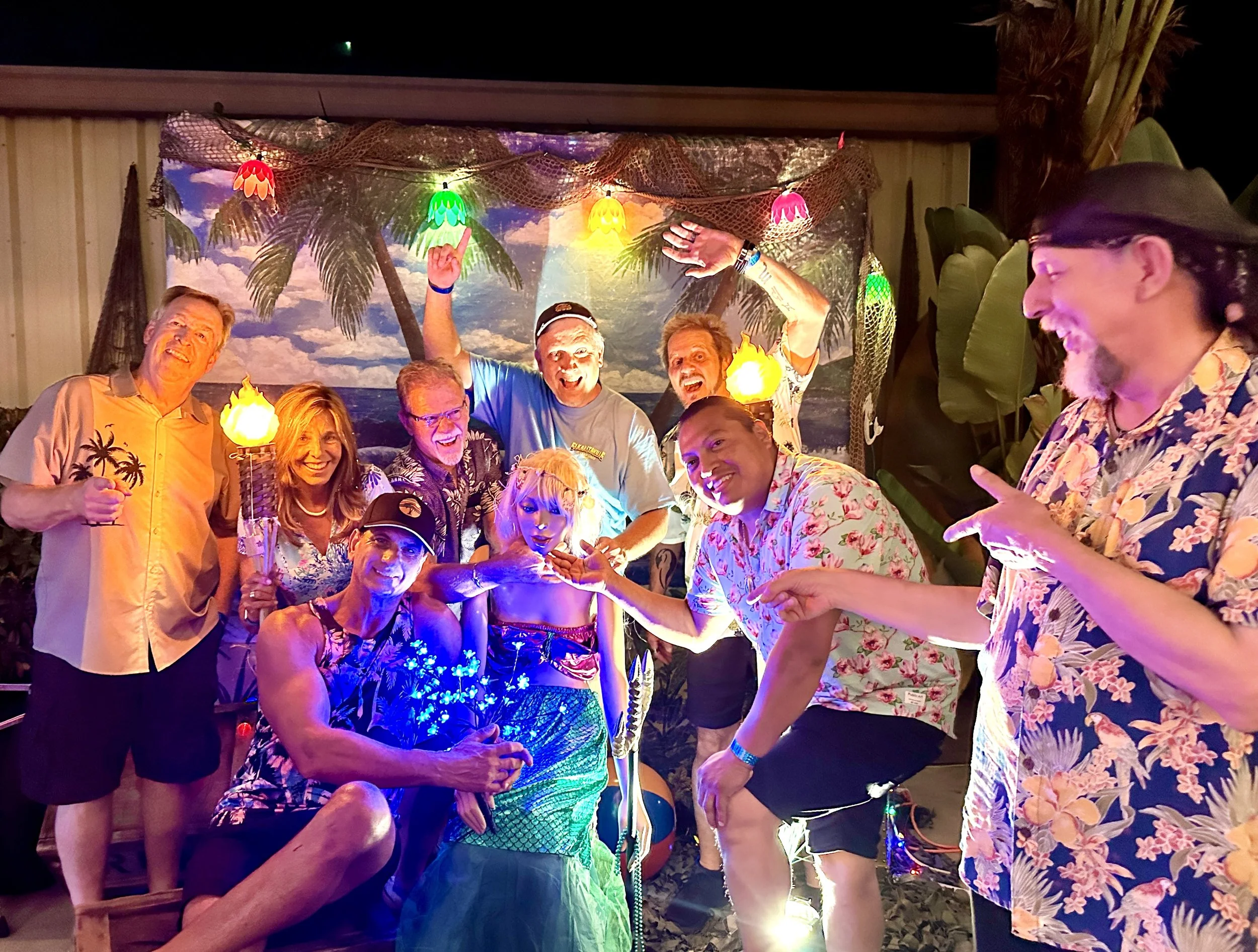 Group of people at a tropical-themed party, smiling and posing with a woman dressed as a mermaid, in front of a beach backdrop with palm trees and sunset, colorful lights and decorations.