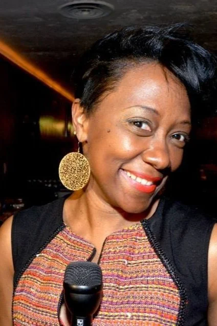African-American woman holding a microphone, wearing a textured dress and large round earrings in a dark setting.