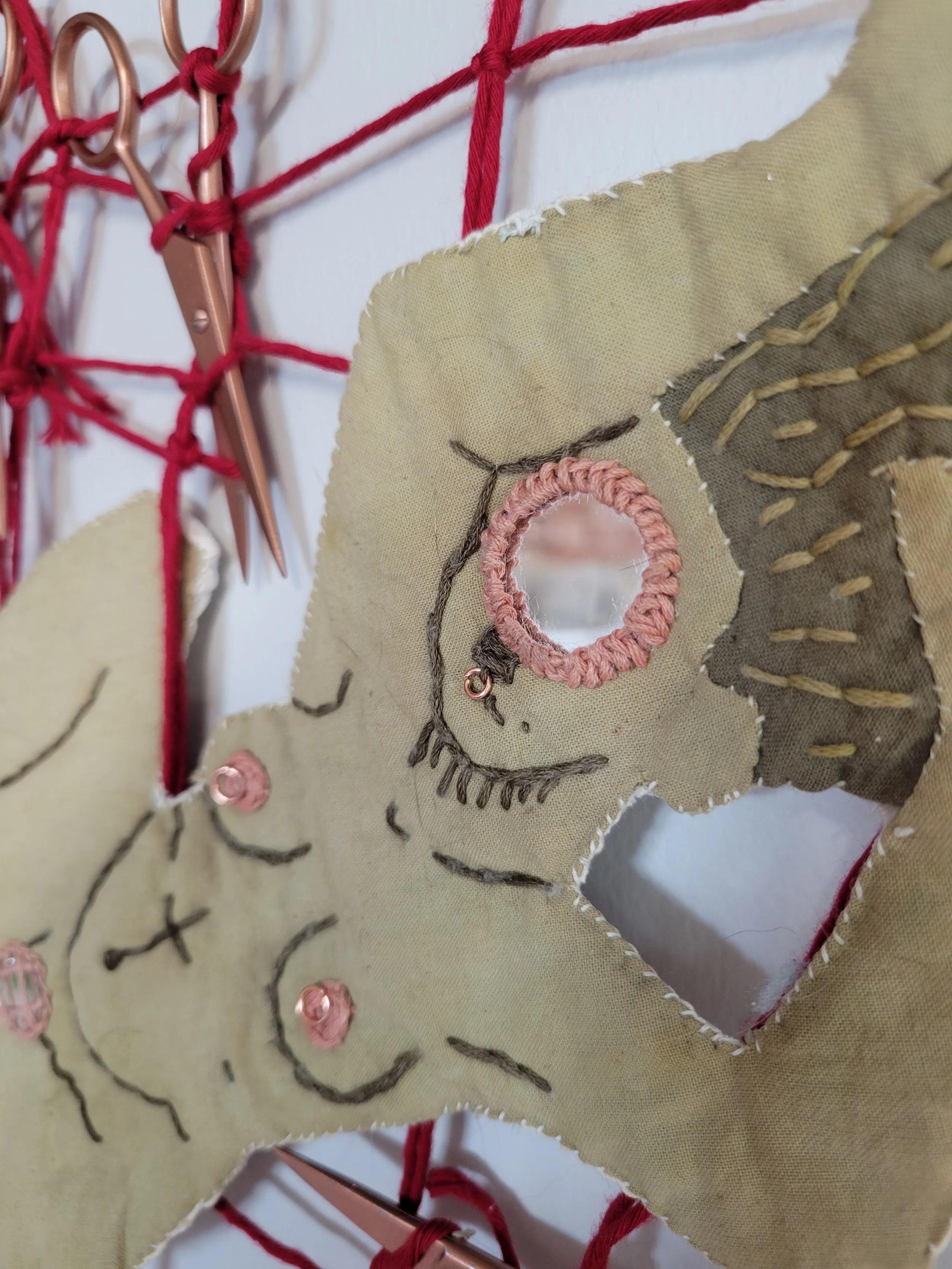 Close-up of a fabric wall hanging with embroidered face and decorative elements, hung on a red rope with copper-colored clips.