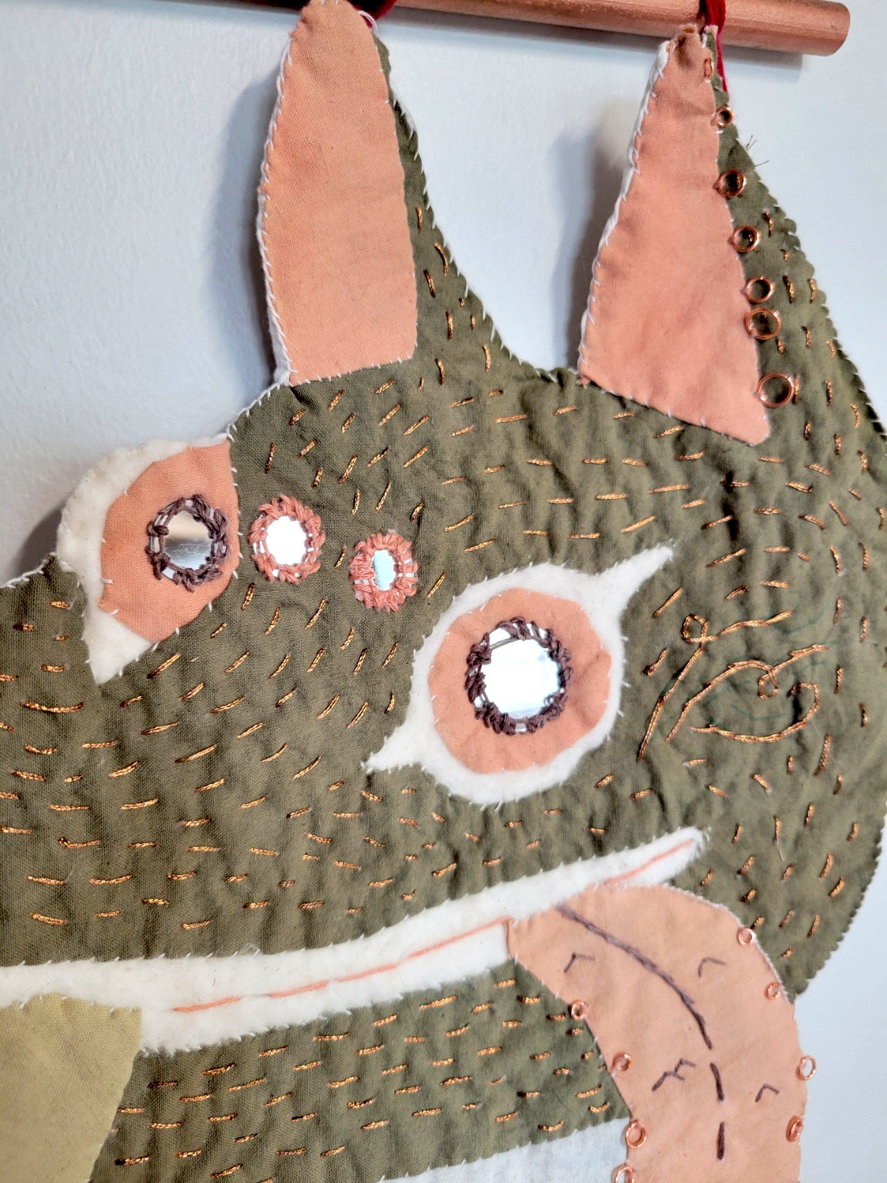 Close-up of a fabric decoration resembling a cartoonish animal face with stitched details, pink ears, and metallic embellishments.