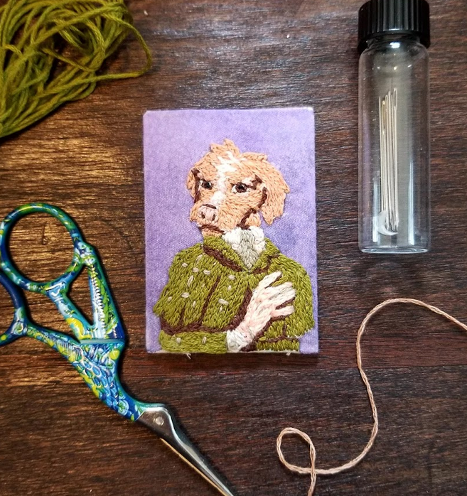 Embroidery kit with a needle, thread, scissors, a small embroidered portrait of an anthropomorphic dog wearing a green jacket on purple fabric, and some green yarn on a wooden surface.