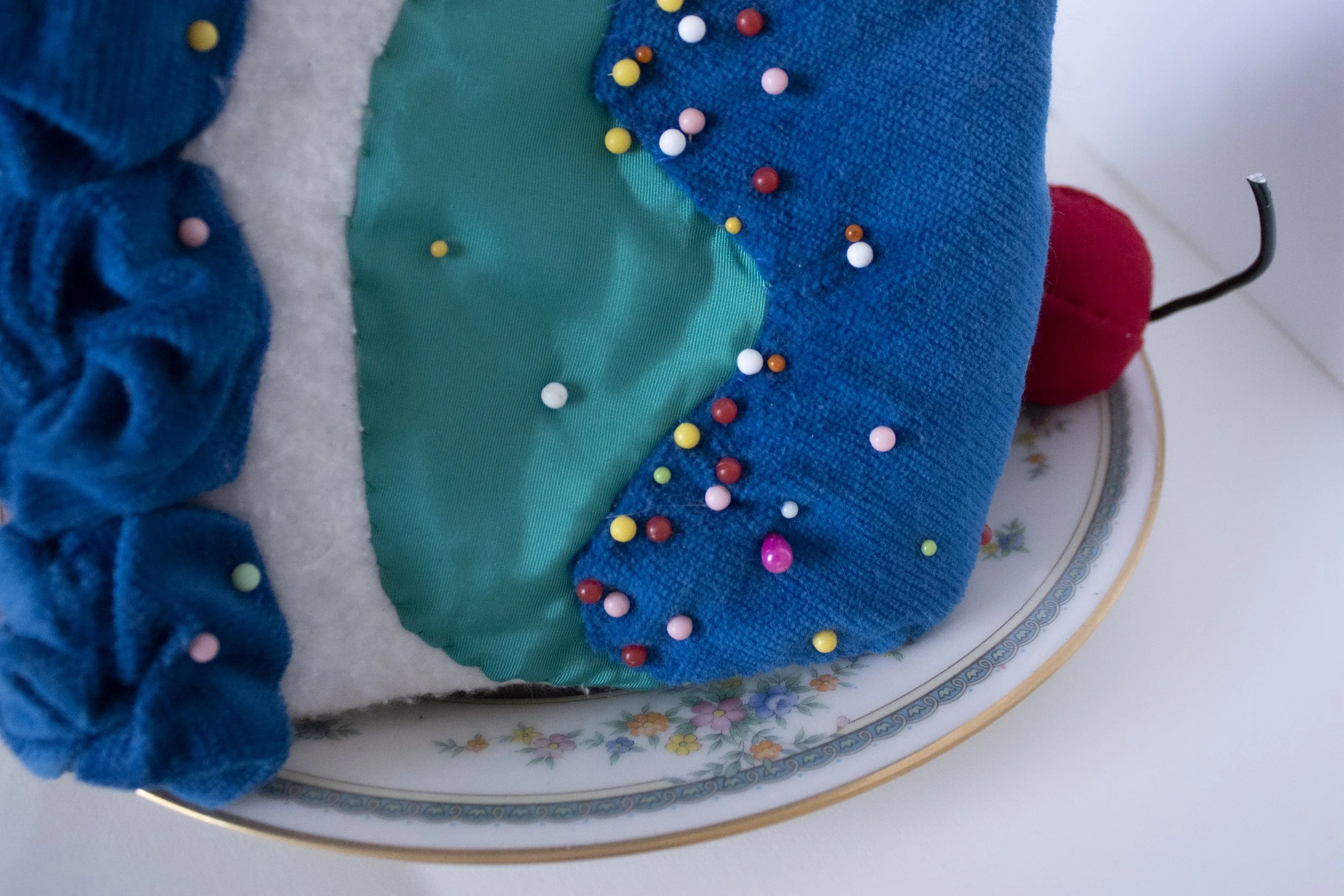 Close-up of a colorful fabric item with a patch of teal satin, decorated with multicolored pins, on a floral-patterned round plate.
