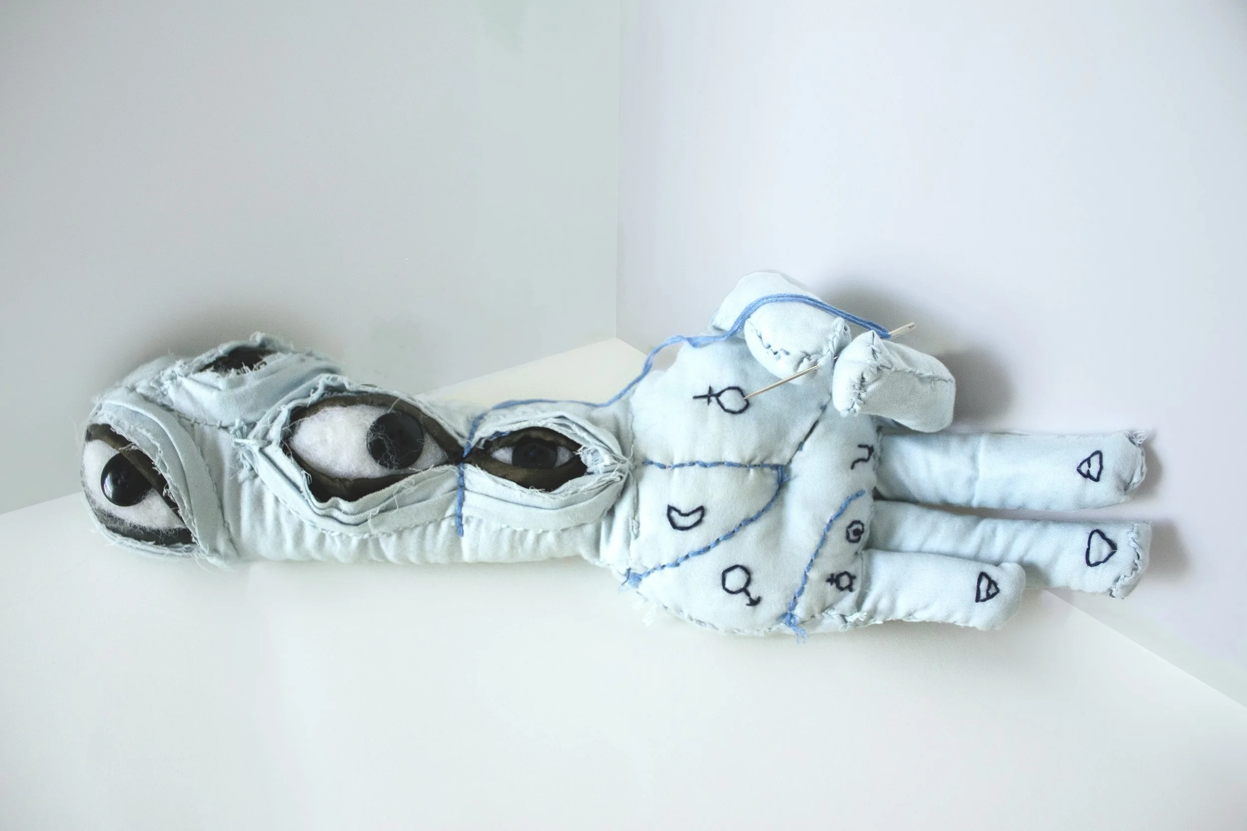 A handmade art doll with stitched fabric body, large black eyes, and blue thread detailing, lying on a white surface in a corner.