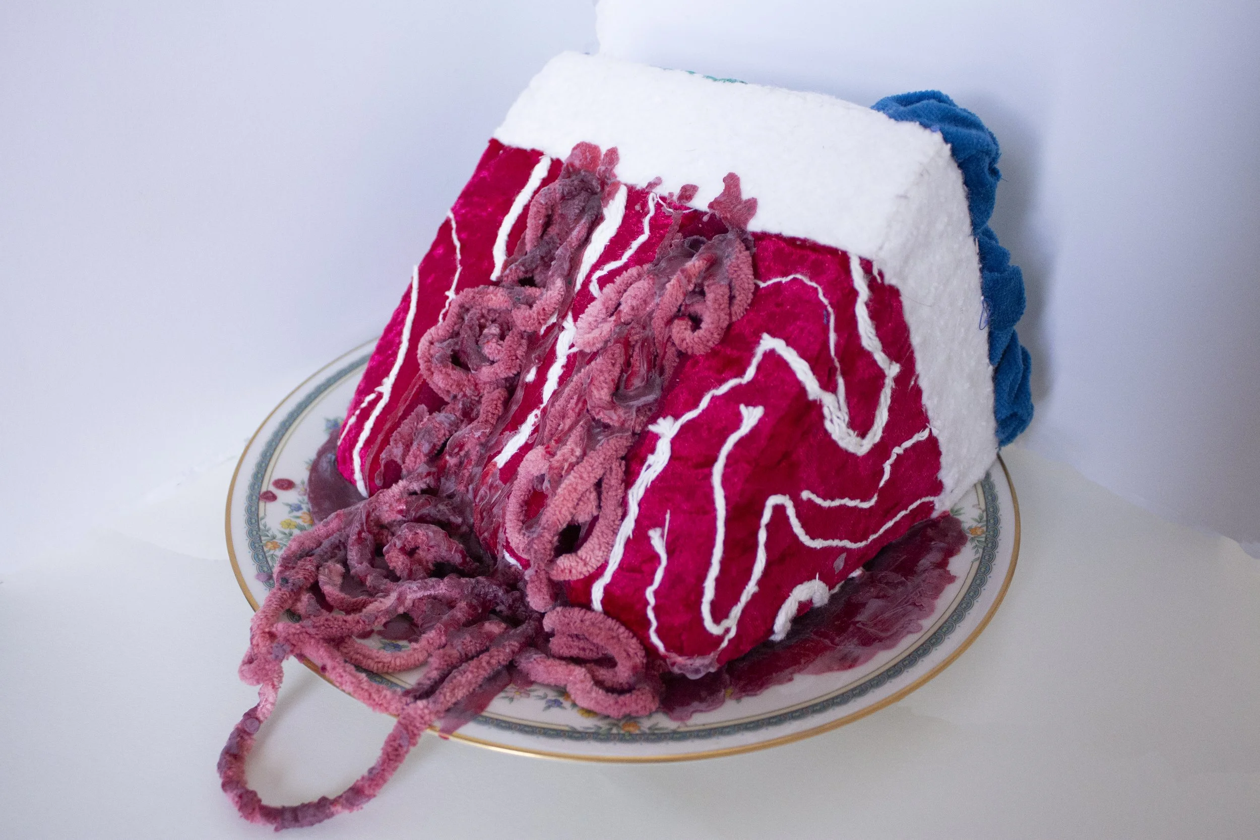 A plate with a large, colorful, plush meat cake textile sculpture with red velvet, white batting, and blue scrap fabric. Pink yarn that looks like guts spills off the plate.